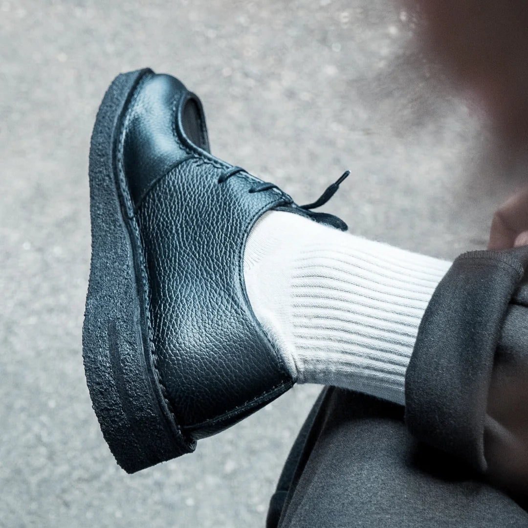 Nahaufnahme des Fußes einer Person, die einen schwarzen Lederschuh mit schwarzen Schnürsenkeln, weiße Rippensocken und eine dunkle Hose trägt und auf dem gegenüberliegenden Knie auf einem hellgrauen, strukturierten Boden ruht.