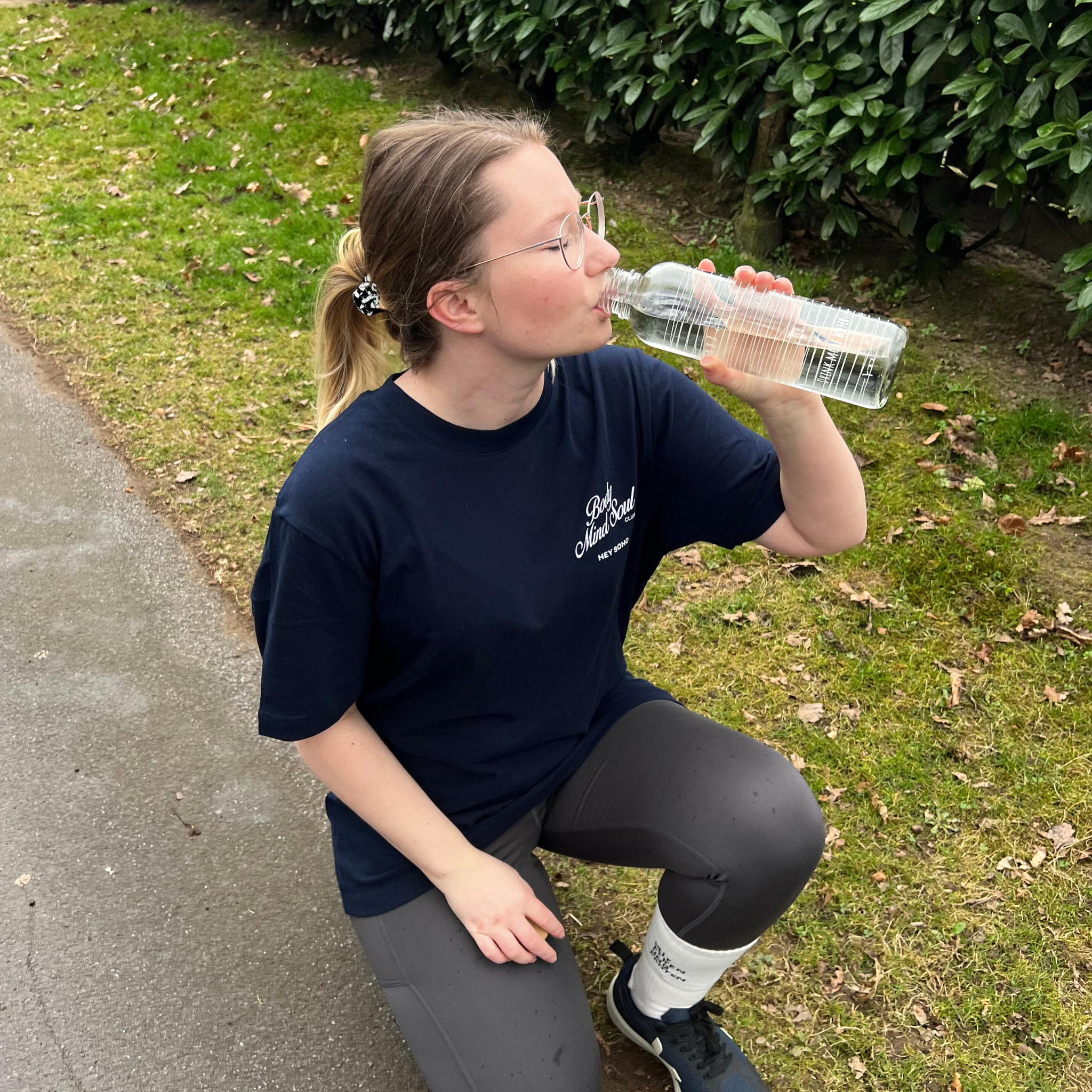 Eine Person in sportlicher Kleidung kniet auf einem Gehweg und trinkt aus einer Wasserflasche, umgeben von Grünflächen.