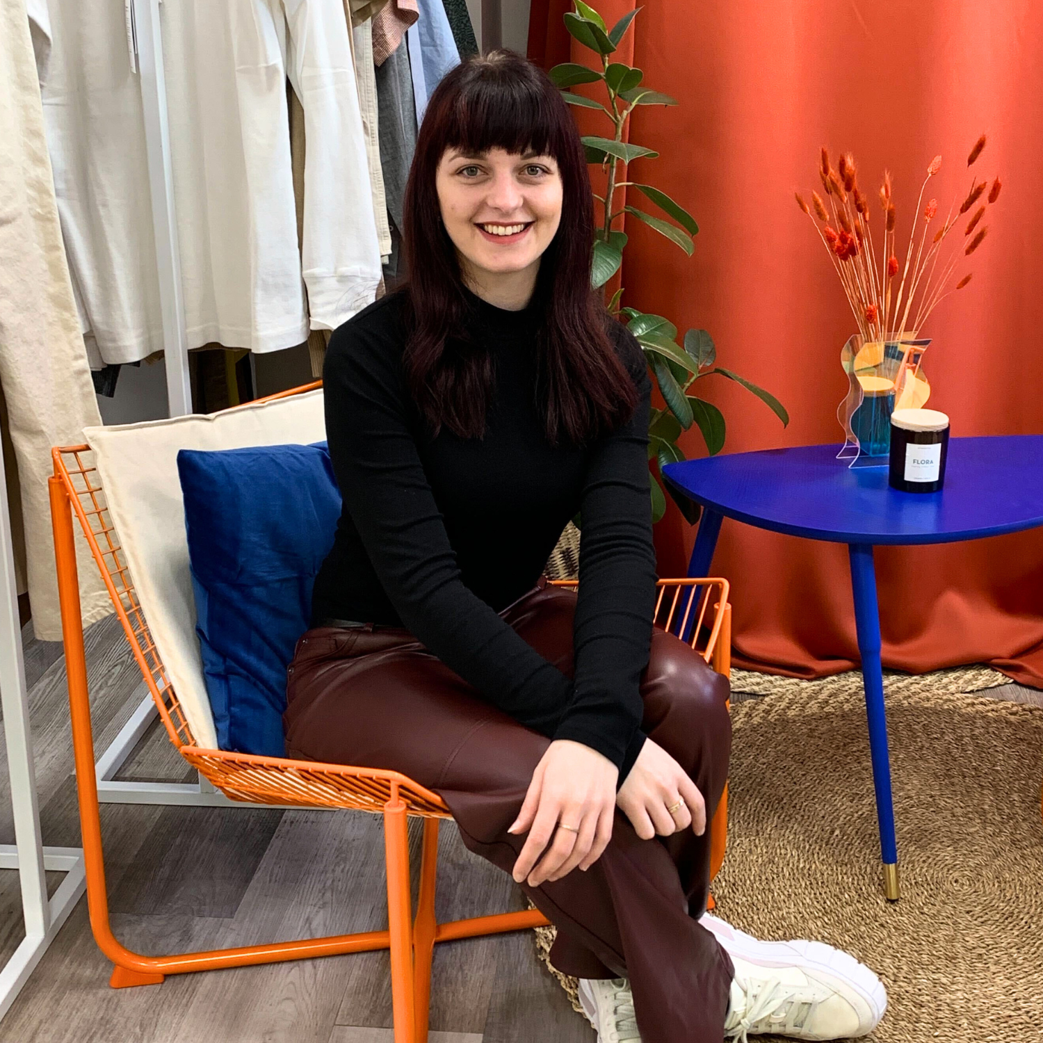 Eine Frau sitzt auf einem orangefarbenen Stuhl, umgeben von Kleidung, einer Pflanze und einem blauen Tisch mit Kerze.