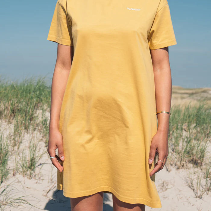 Eine Person in einem kurzärmeligen gelben Kleid steht an einem Sandstrand mit Gras und blauem Himmel im Hintergrund. Ihr Gesicht ist nicht zu sehen, und sie trägt Ringe und ein Armband.