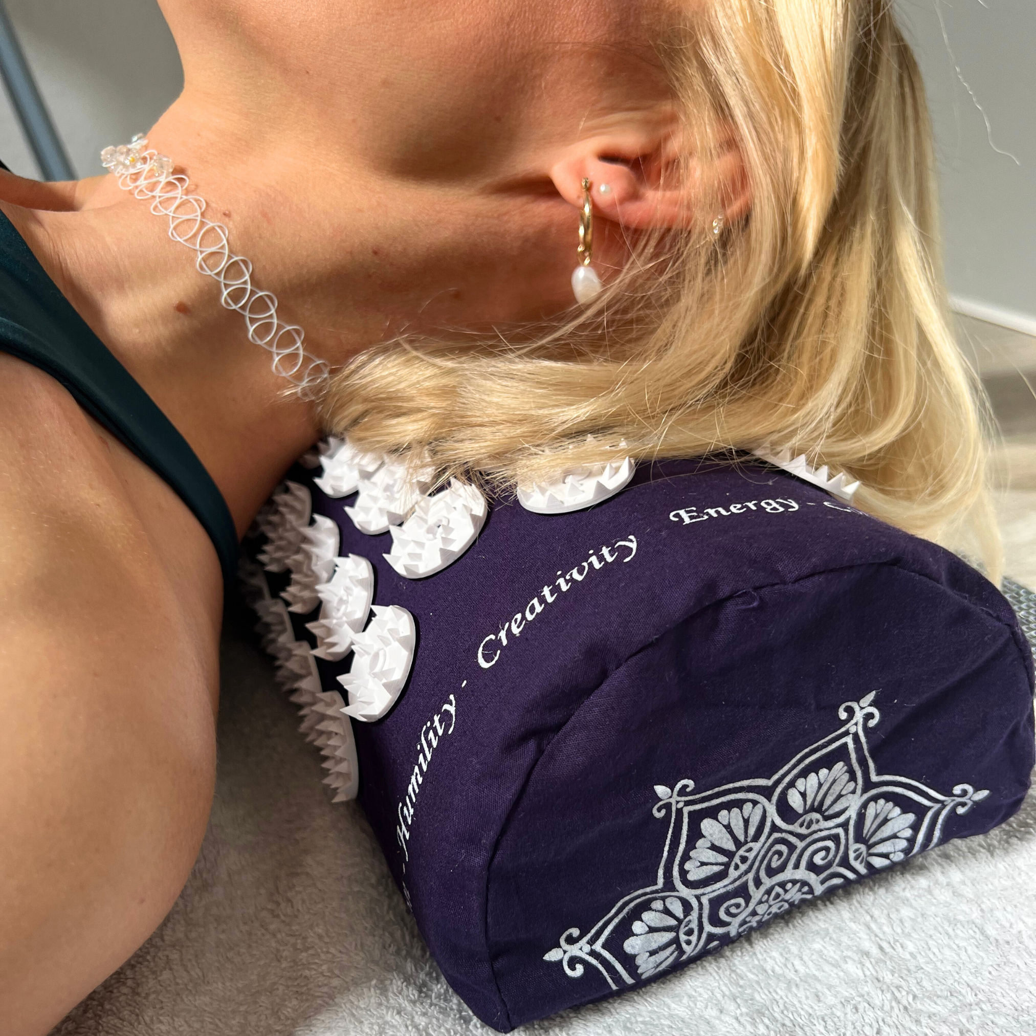 Frau liegt mit dem Nacken auf einem Akupressur-Kissen mit weißen Spitzen und trägt ein transparentes Halsband.