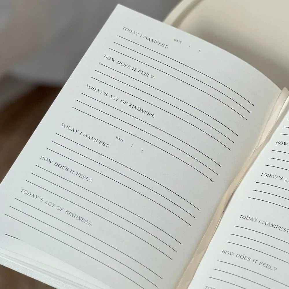 Ein offenes Tagebuch mit Aufforderungen auf jeder Seite, einschließlich Today I Manifest, How does it feel? und Todays Act of Kindness, jeweils mit Zeilen darunter zum Schreiben von Antworten. Das Tagebuch liegt auf einer weißen Unterlage.