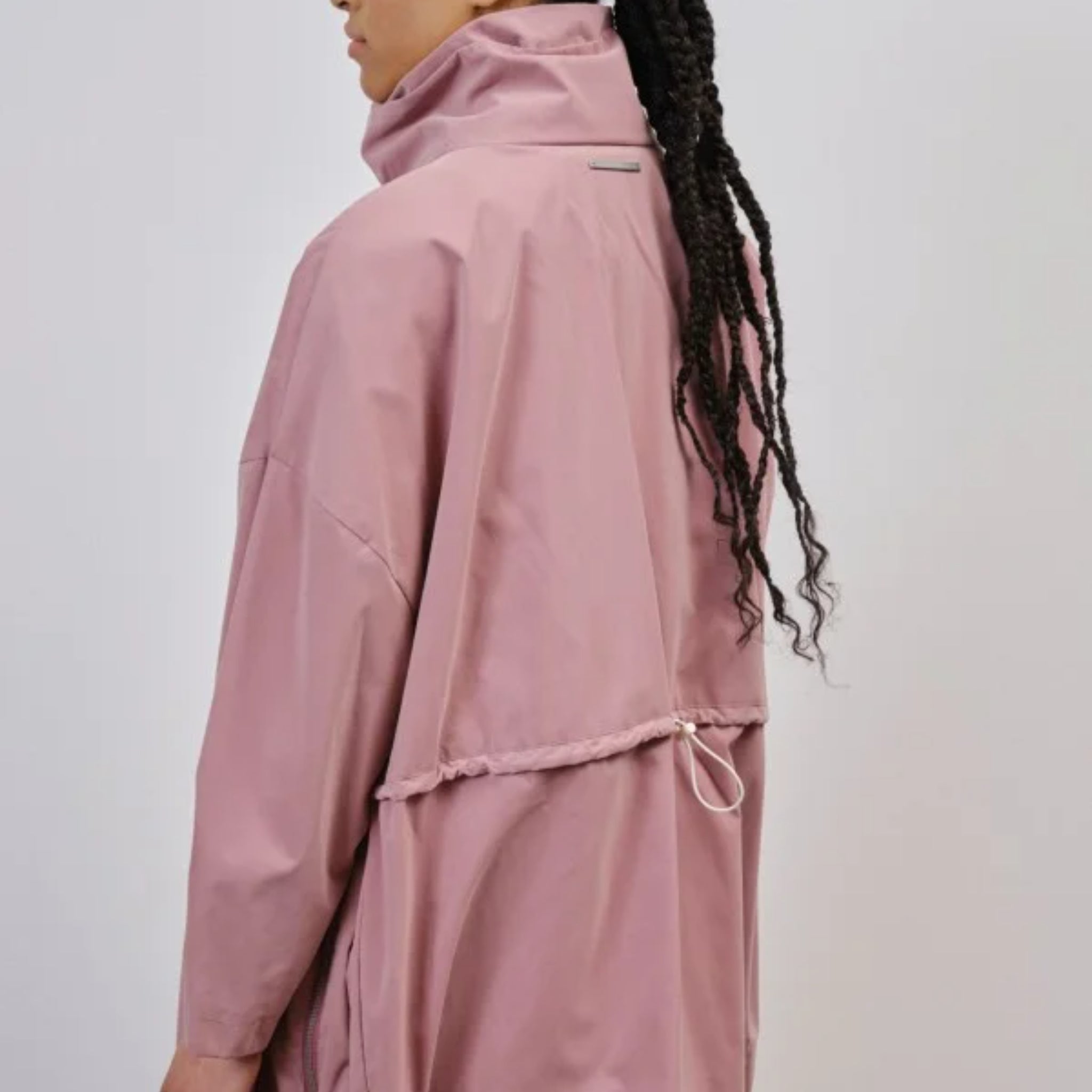 Person in a pink jacket with braided hair, viewed from the back, standing against a plain background.