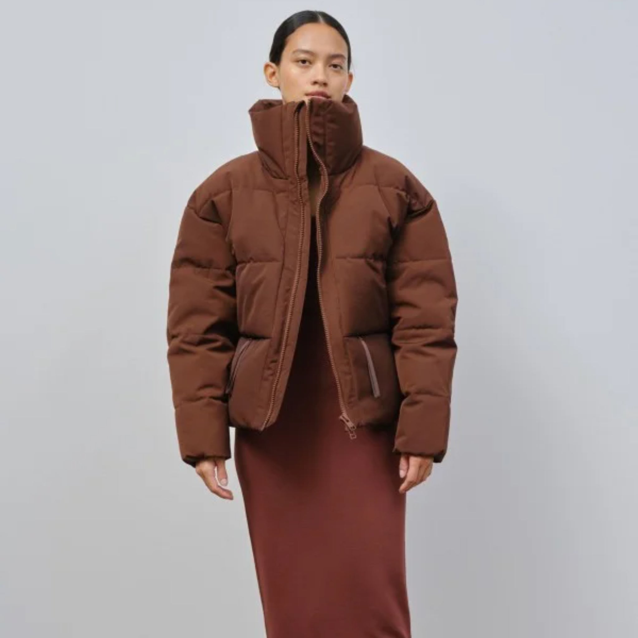 Person in brauner Steppjacke und langem braunen Kleid steht vor einem grauen Hintergrund.