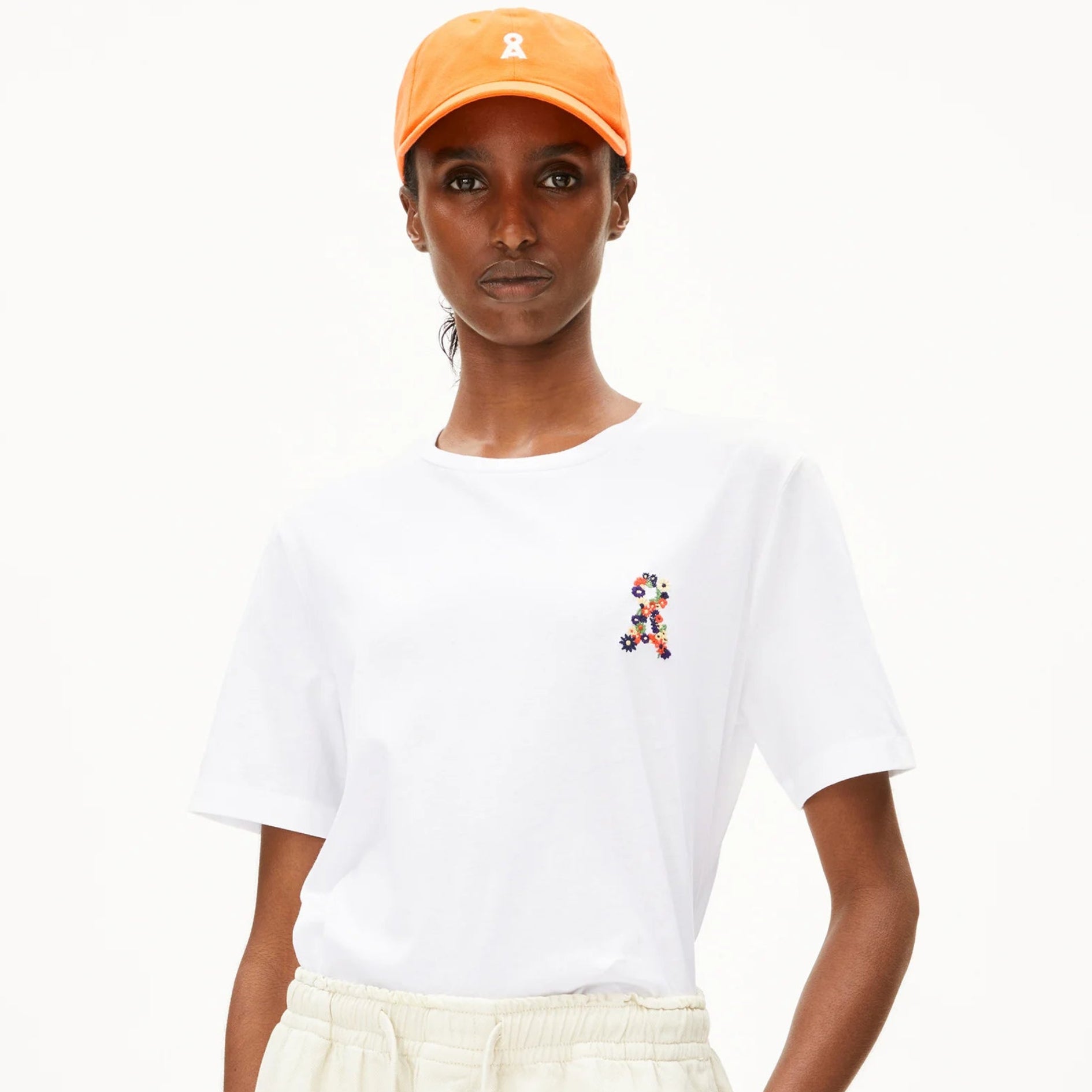 Eine Person, die ein weißes T-Shirt mit einem kleinen bunten Muster, eine cremefarbene Hose und eine orangefarbene Mütze trägt, steht mit den Händen in den Taschen vor einem schlichten weißen Hintergrund.