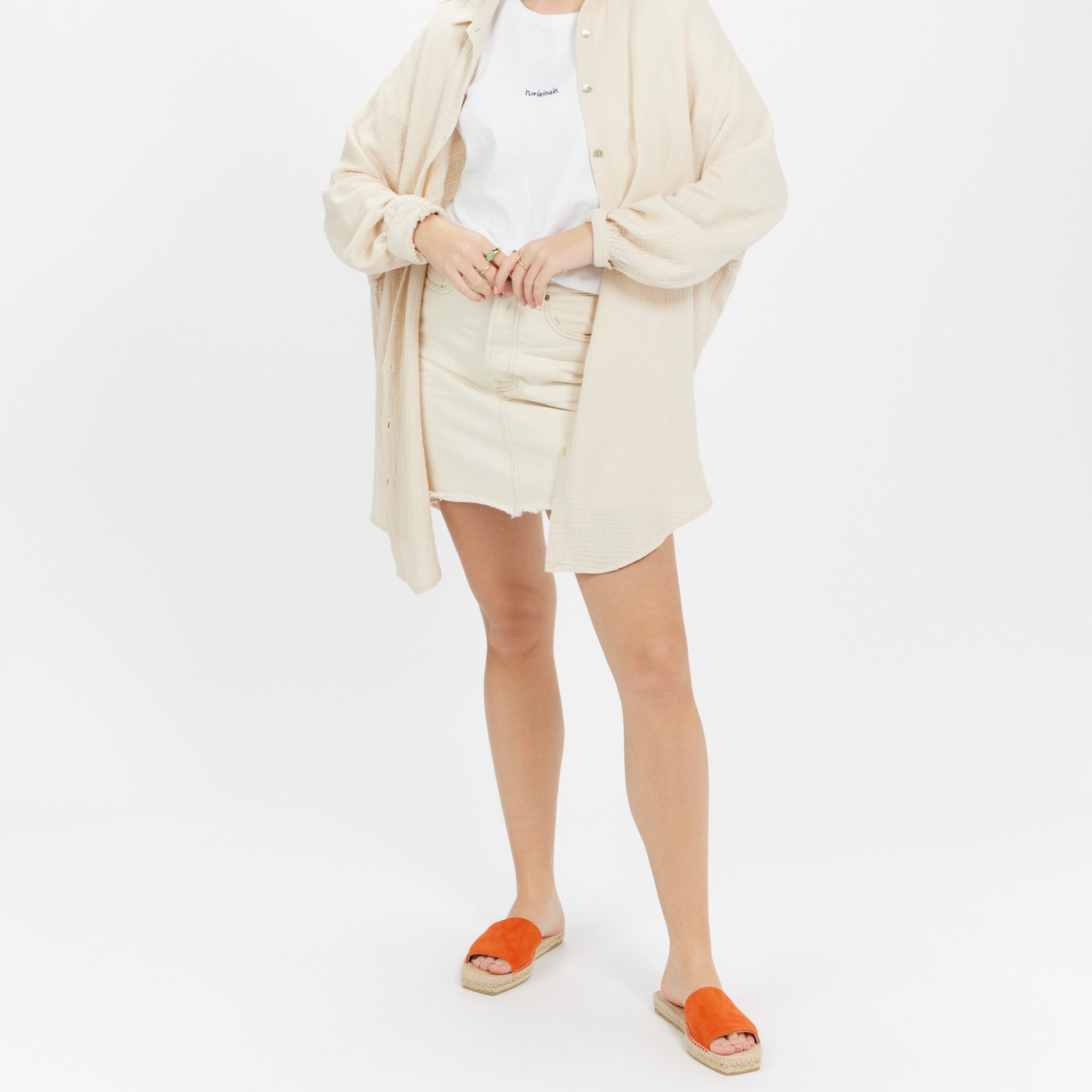 Person in beiger Bluse, weißem T-Shirt, hellem Rock und orangefarbenen Sandalen vor weißem Hintergrund.