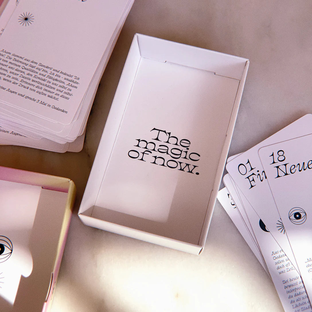 Eine weiße Schachtel mit der Aufschrift The magic of now. im Inneren, umgeben von verstreuten Karten mit Text und einfachen Augenillustrationen auf einer hellen Oberfläche.