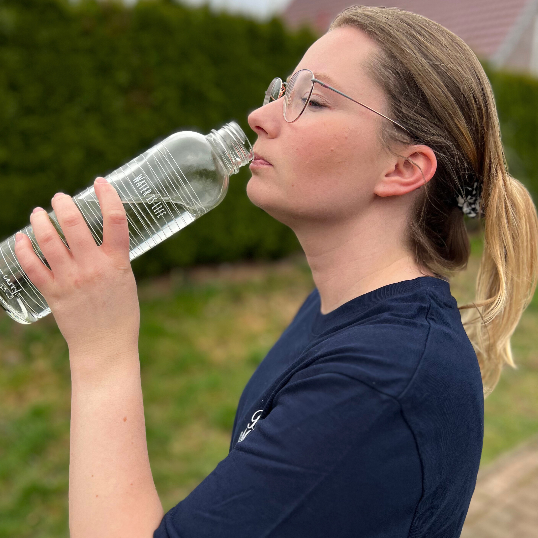 Eine Person mit Brille trinkt aus einer Wasserflasche im Freien.