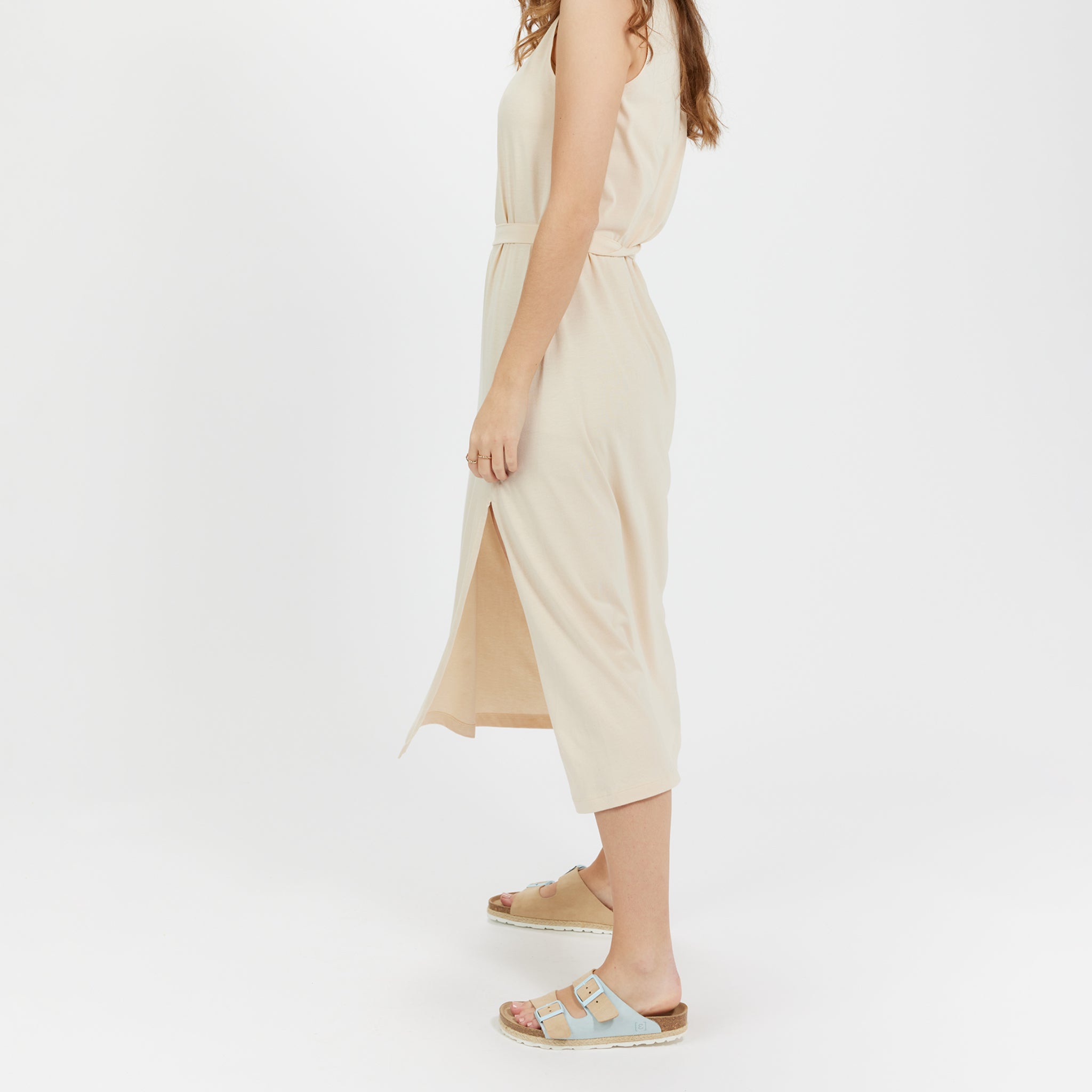 Eine Frau trägt ein beigefarbenes Kleid und Sandalen, stehend vor einem weißen Hintergrund.