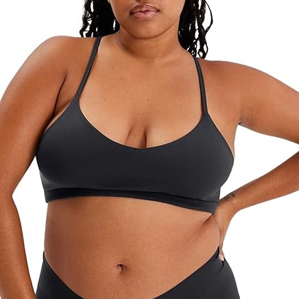 Eine Frau mit langen geflochtenen Haaren trägt einen einfachen schwarzen Bralette und eine passende schwarze Bikinihose. Sie steht mit entspanntem, selbstbewusstem Gesichtsausdruck vor der Kamera und stützt ihre Hand auf ihre Hüfte.