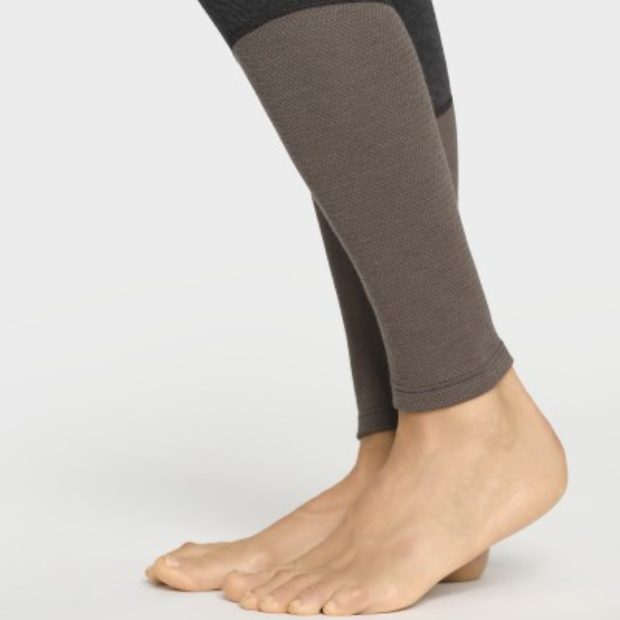 Nahaufnahme von nackten Füßen und Unterschenkeln in dunklen, eng anliegenden Leggings mit strukturierten braunen Stoffbahnen an den Waden. Der Hintergrund ist einfarbig weiß.