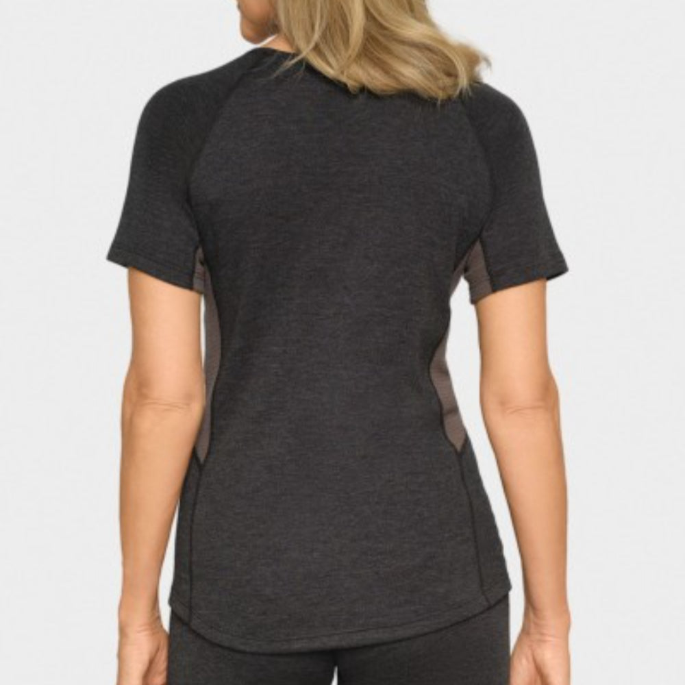 Eine Frau mit blondem Haar ist von hinten zu sehen. Sie trägt ein kurzärmeliges, dunkelgraues Sporthemd und eine passende Hose vor einem schlichten, hellen Hintergrund.