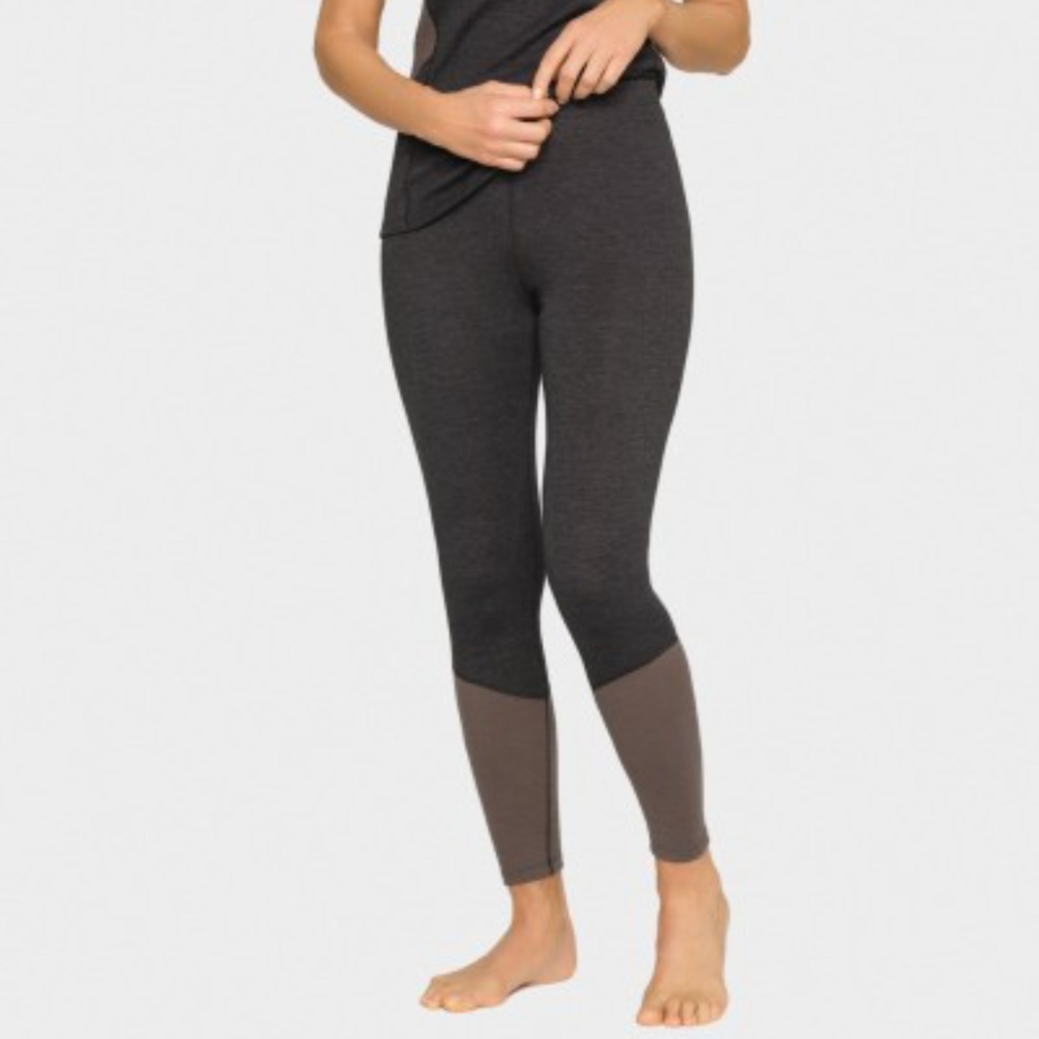 Eine Person steht barfuß vor einem schlichten Hintergrund und trägt eine dunkelgraue Leggings mit einem helleren grauen Abschnitt an den Unterschenkeln und ein dazu passendes dunkelgraues Oberteil. Ihre Hände passen den Bund an.