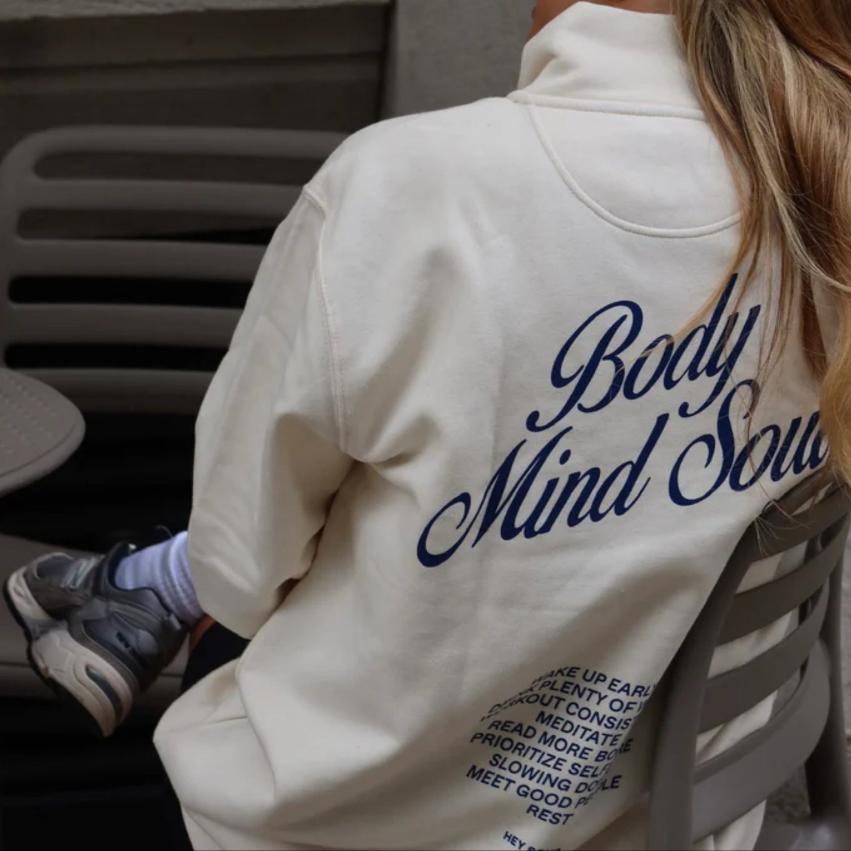 Person in einem weißen Pullover mit dem Text "Body Mind Soul" und mehreren Tipps darauf, sitzt auf einem Stuhl.
