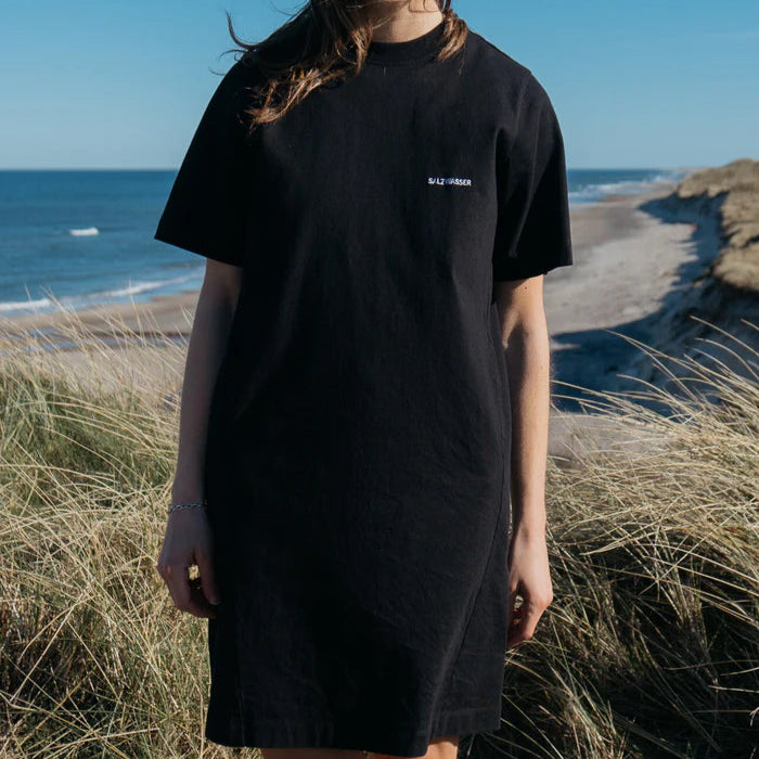 Eine Person, die ein schwarzes T-Shirt trägt, steht im hohen Strandgras, während im Hintergrund das Meer und die sandige Küste unter einem klaren blauen Himmel zu sehen sind.