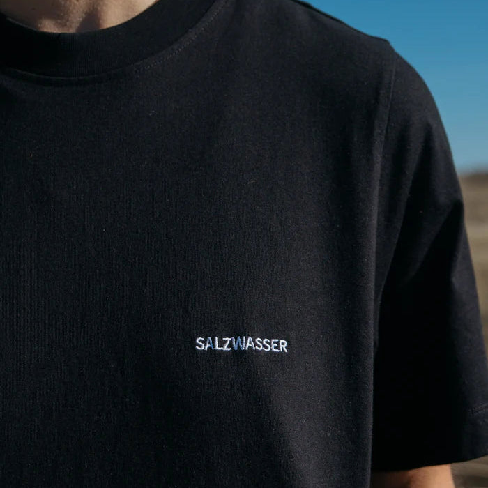 Eine Person, die ein schlichtes schwarzes T-Shirt trägt, auf dessen Brust in weißer Farbe das Wort SALZWASSER aufgestickt ist, steht im Freien unter einem klaren blauen Himmel.