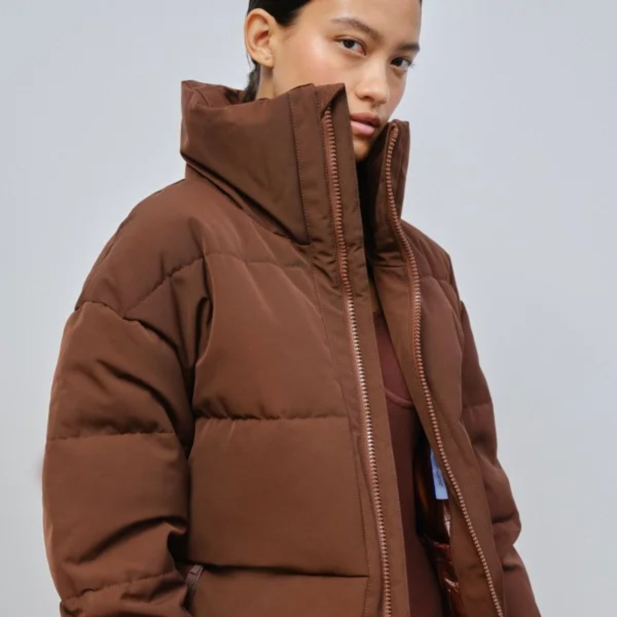 Person in a brown puffer jacket with a high collar looking at the camera against a gray background.