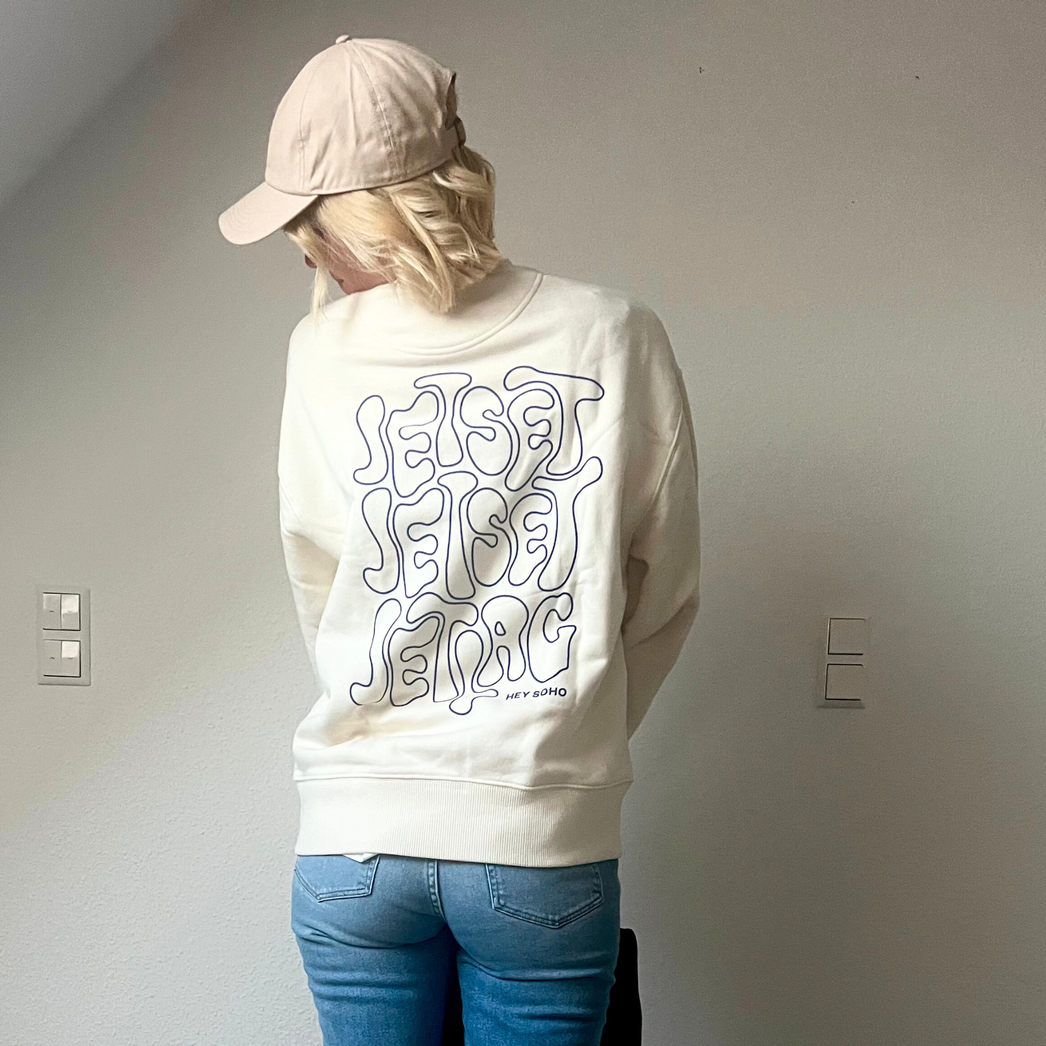 Person mit blonden Haaren und Baseballkappe trägt einen cremefarbenen Pullover mit dem Schriftzug "Jetset Jetlag".