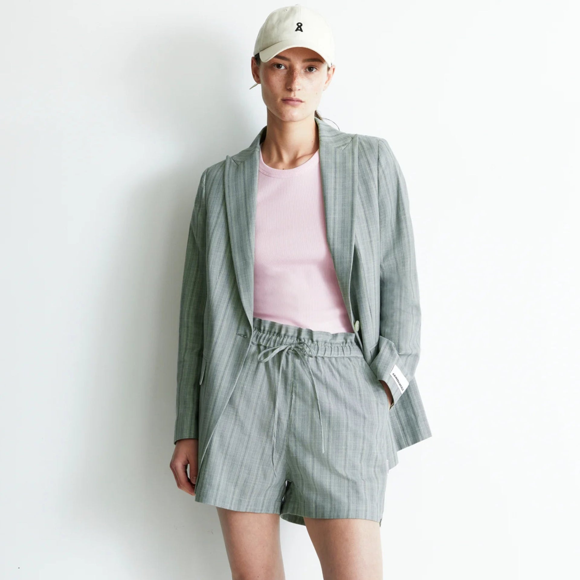 Eine Person steht vor einer weißen Wand und trägt einen hellgrauen Nadelstreifenblazer und dazu passende Shorts, ein rosa T-Shirt, weiße Turnschuhe und eine weiße Baseballmütze.