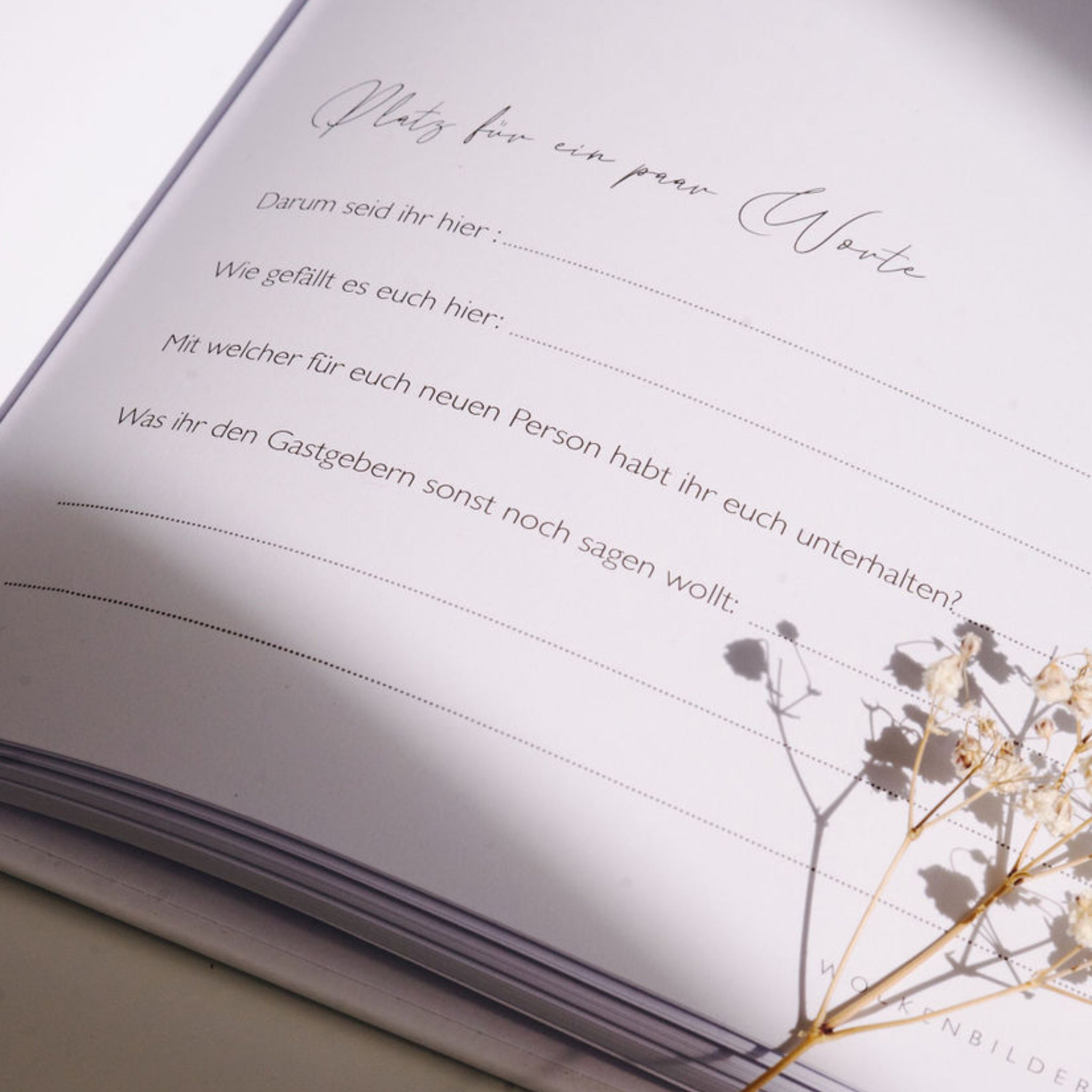 Aufgeschlagenes Gästebuch mit Fragen für Einträge, dekoriert mit einem Zweig getrockneter Blumen.