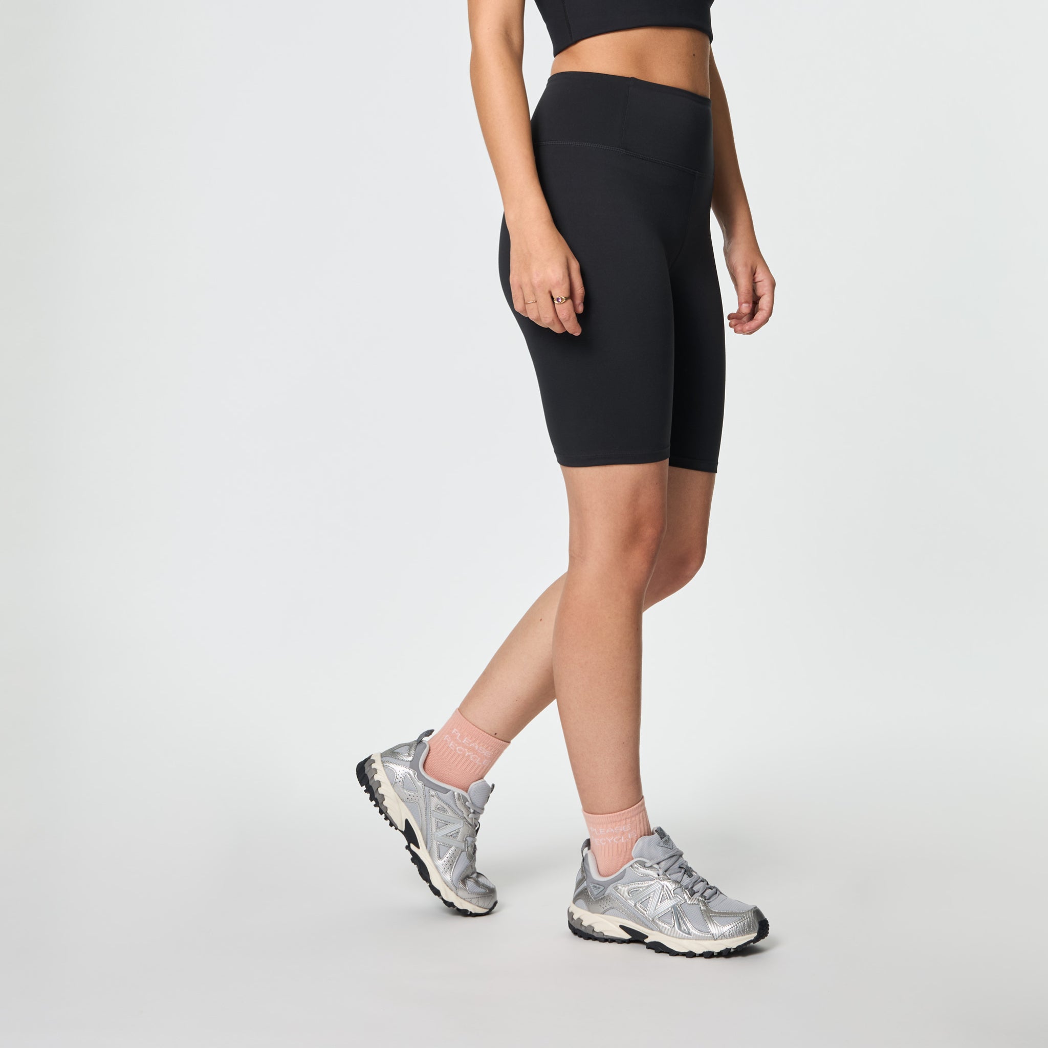 Frau in schwarzer Sportbekleidung, Fahrradshorts und grauen Turnschuhen vor weißem Hintergrund.