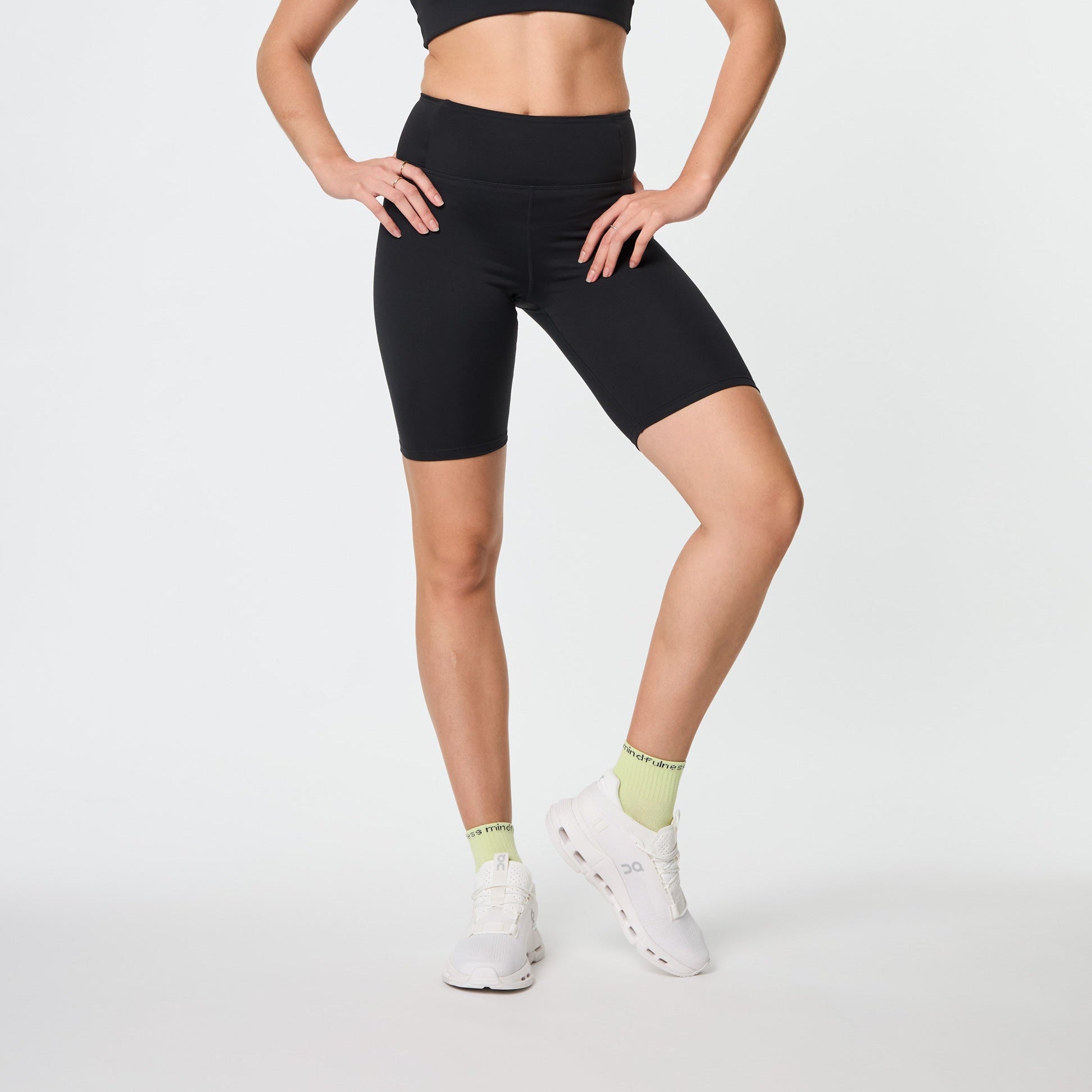 Frau in schwarzem Sport-BH und Shorts, grünen Socken und weißen Turnschuhen posiert vor weißem Hintergrund.