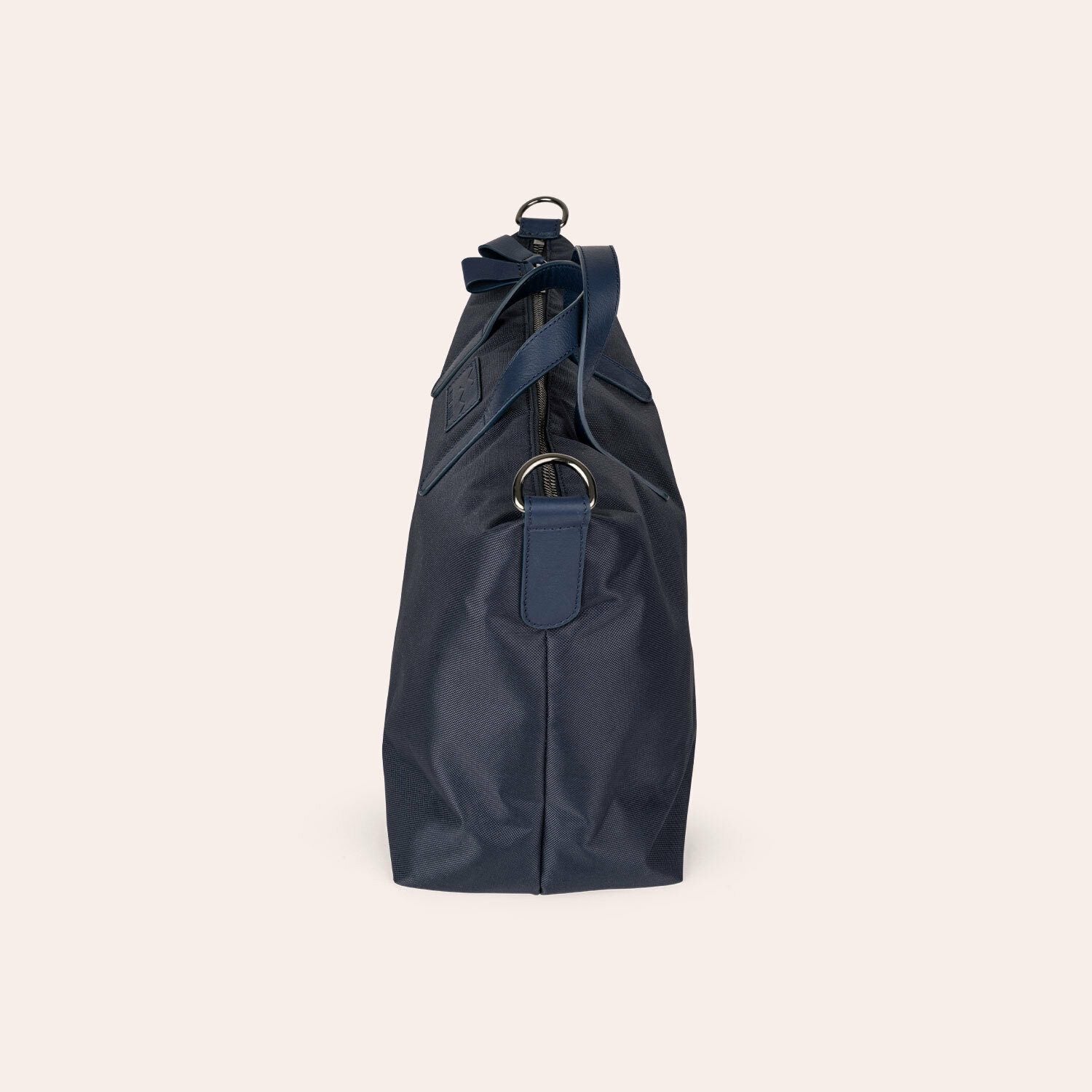 Seitenansicht einer marineblauen Tragetasche mit Ledergriffen und Metallringen vor einem schlichten hellbeigen Hintergrund.