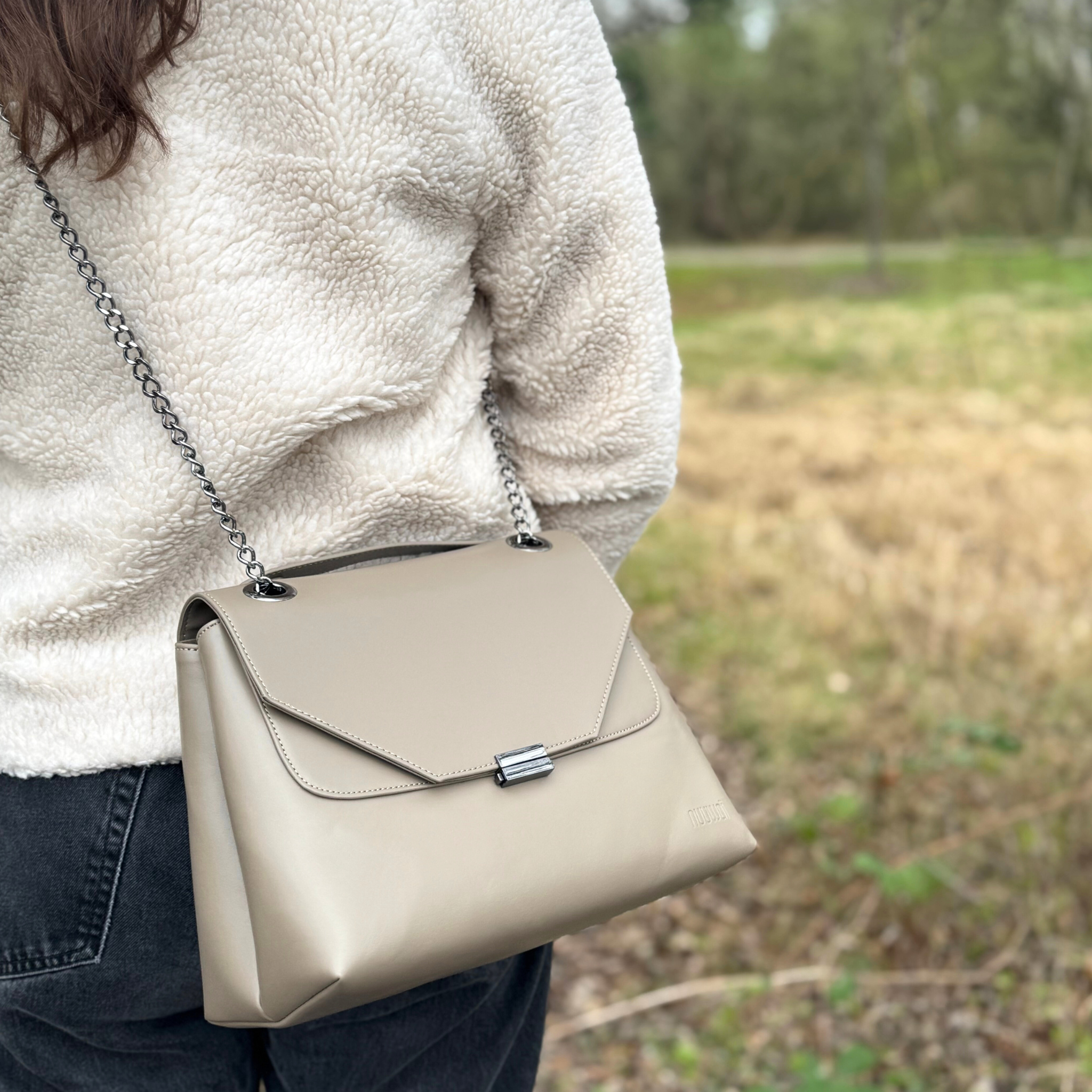 Person mit beigem Mantel trägt eine hellbraune Umhängetasche mit Metallkettenriemen draußen auf einem Feldweg.