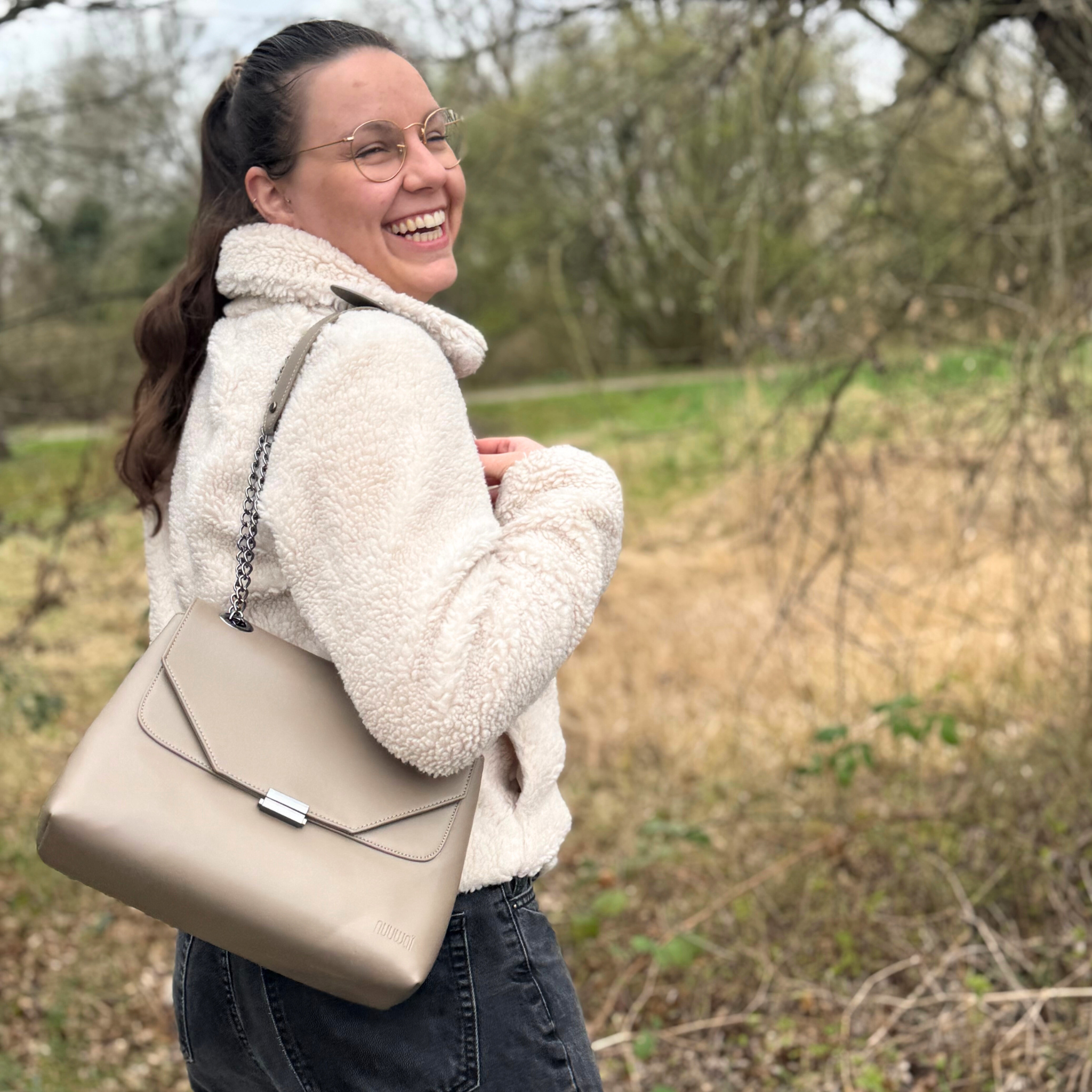 Eine fröhliche Frau in einer weißen Jacke trägt eine beigefarbene Handtasche und steht in einer herbstlichen Landschaft.