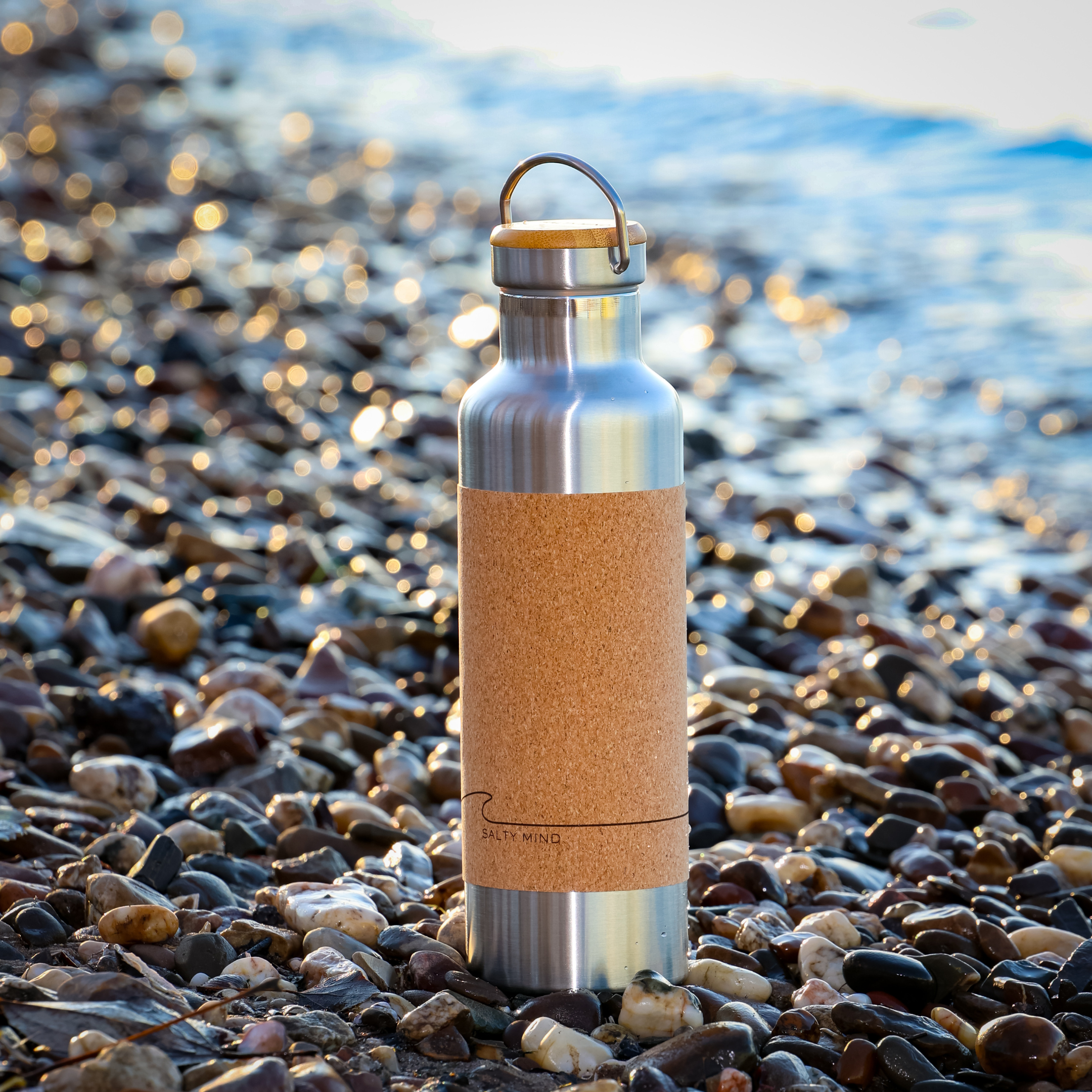 Eine Metallflasche mit Korkverkleidung steht auf einem kieseligen Strand bei hellem Sonnenlicht nahe dem Wasser.