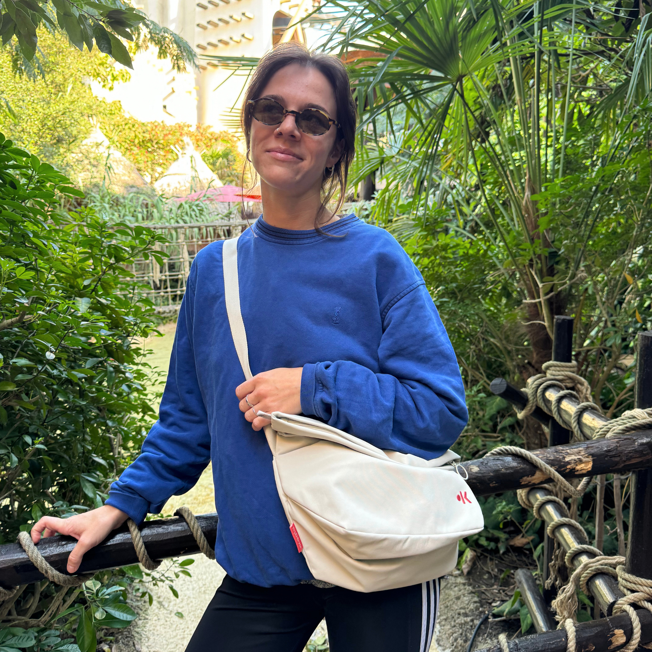 Eine Frau in einem blauen Pullover und Sonnenbrille steht draußen inmitten üppiger Vegetation und lächelt leicht.