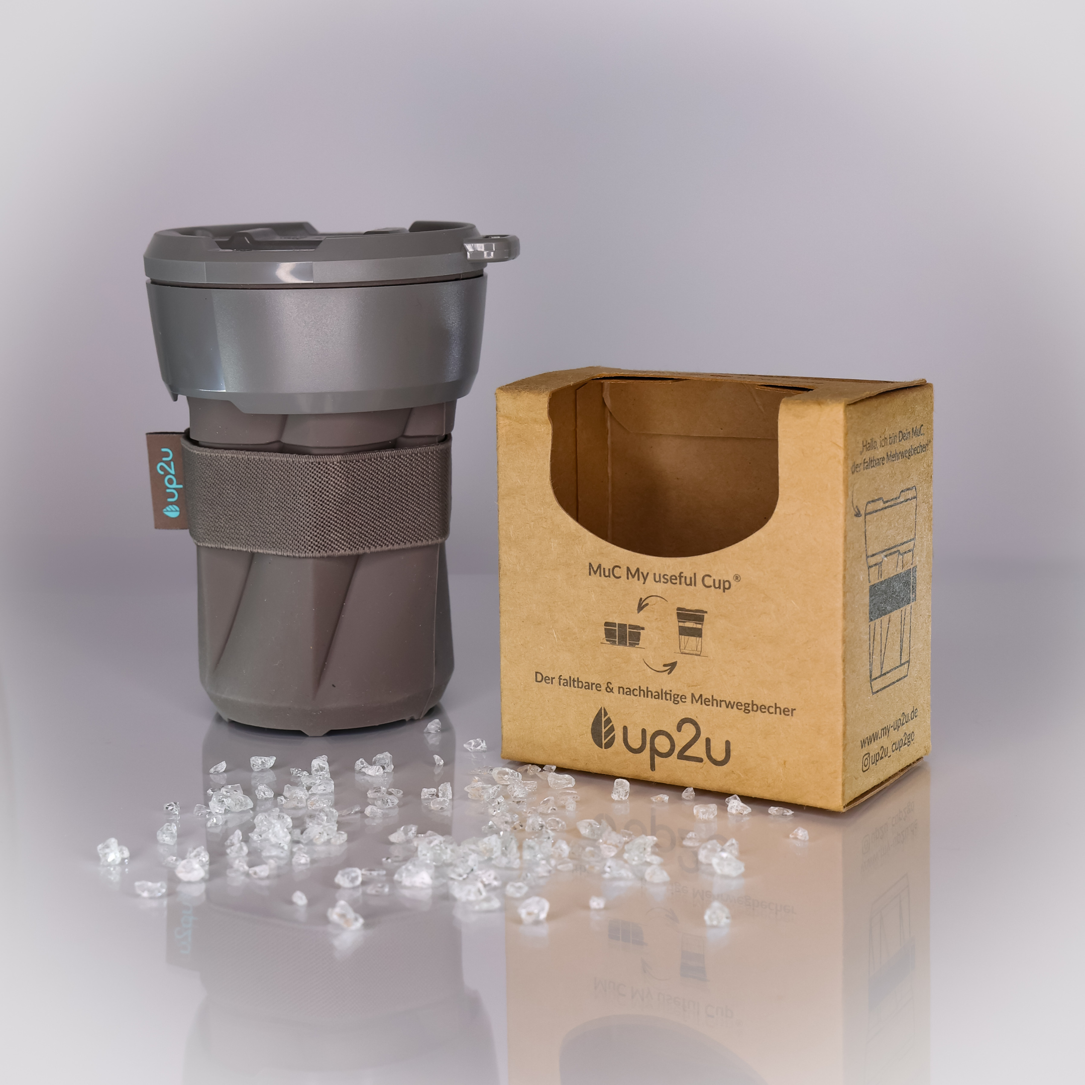Grauer, faltbarer Kaffeebecher mit Kartonverpackung und verstreuten Kristallen auf einer reflektierenden Oberfläche.