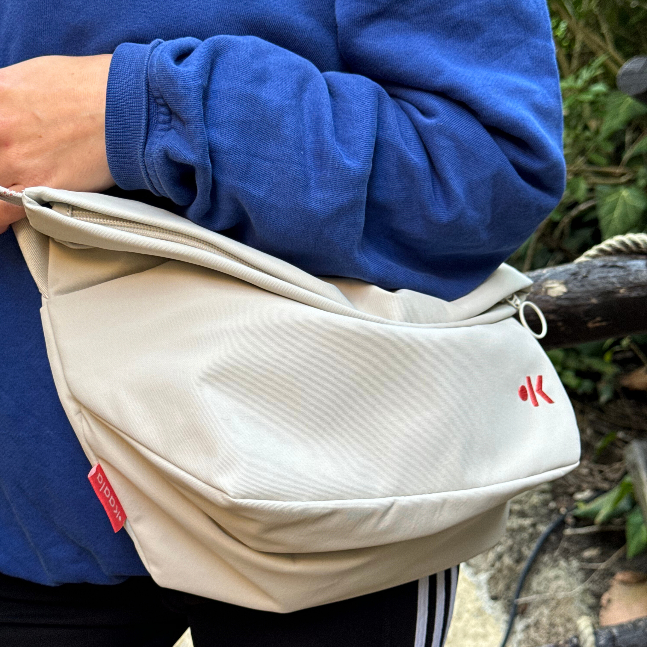 Person in blauem Pullover hält eine cremefarbene Tasche mit rotem Logo und Reißverschluss, draußen im Grünen.