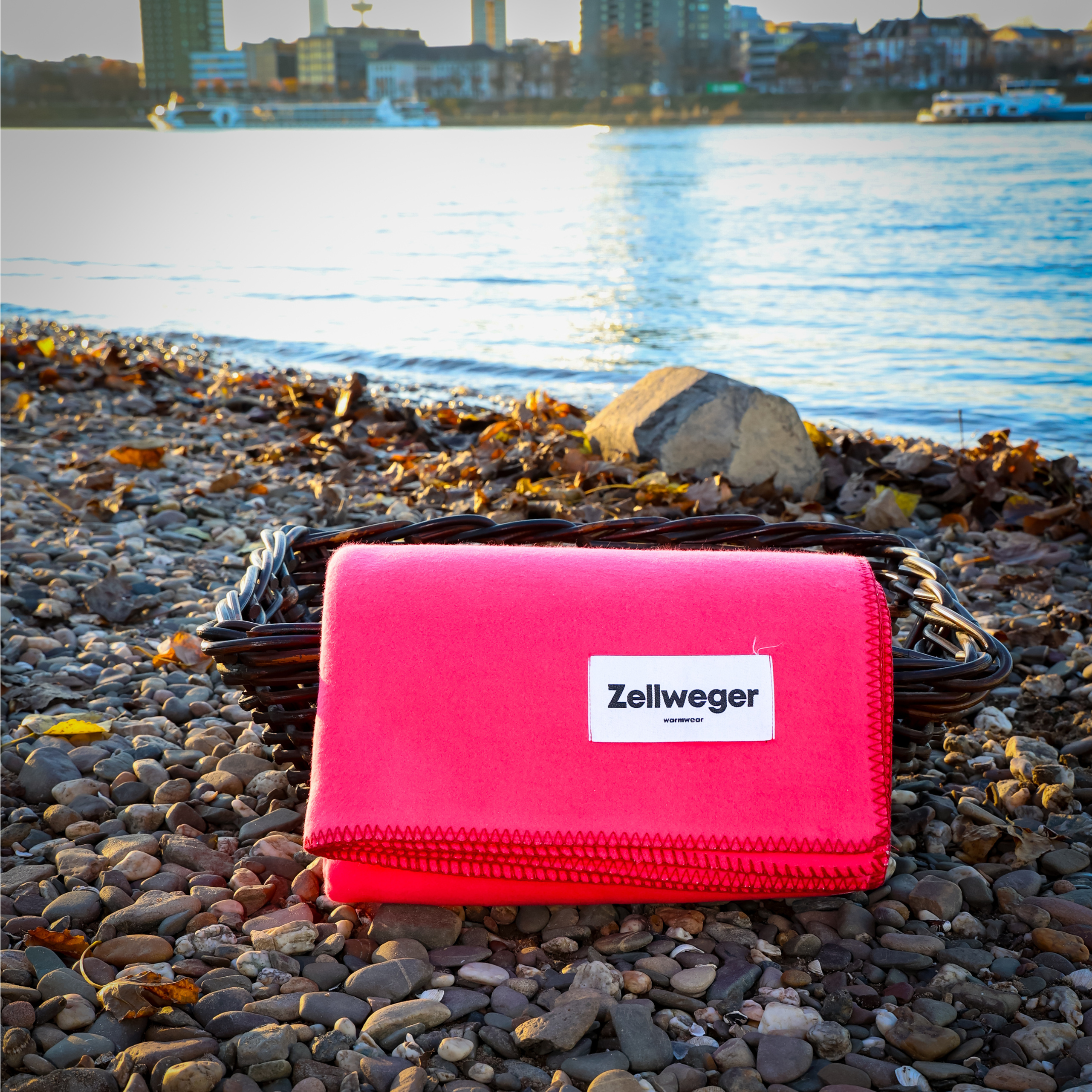 Roter Zellweger-Behälter auf einem Kiesstrand mit Wasser und Stadtsilhouette im Hintergrund.