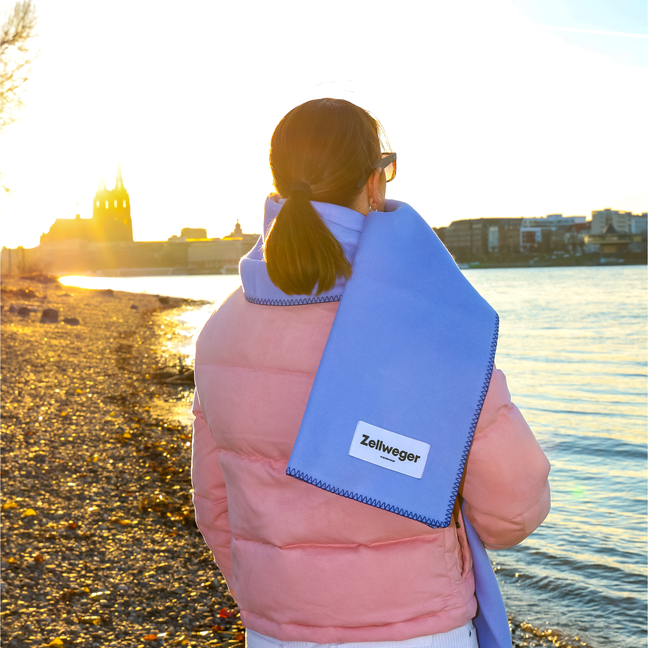 Frau im rosa Mantel mit blauem Schal am Flussufer, im Hintergrund eine Stadtansicht bei Sonnenuntergang.