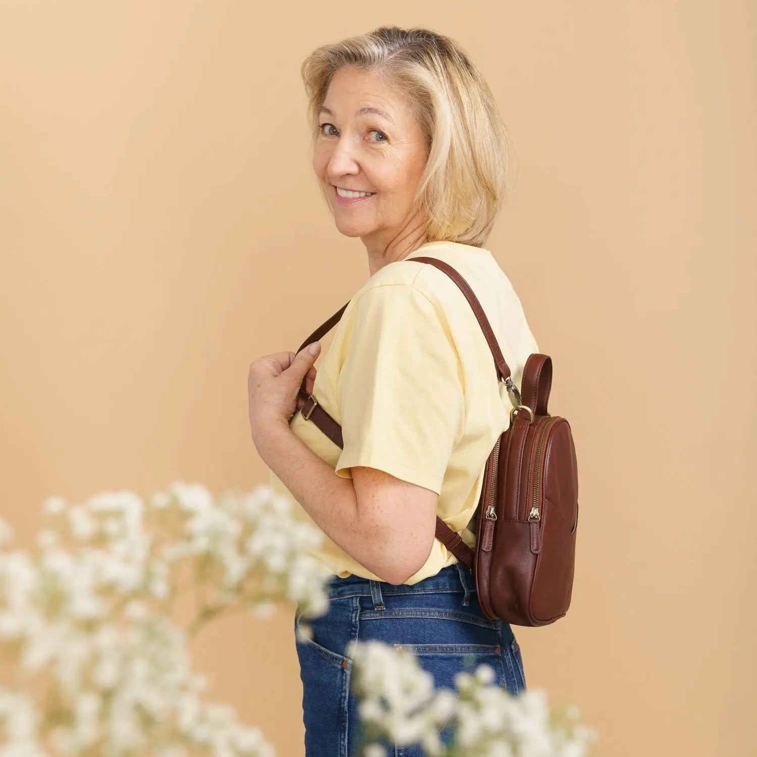 Eine lächelnde ältere Frau mit blondem Haar, die ein gelbes Hemd und blaue Jeans trägt, steht seitlich mit einem braunen Rucksack vor einem beigen Hintergrund. Weiße Blumen sind im Vordergrund unscharf.