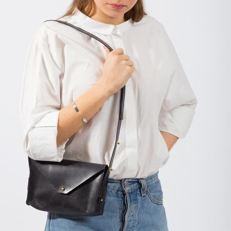 Eine Frau, die ein weißes Button-up-Hemd und blaue Jeans trägt, hält eine schwarze Ledertasche mit Überschlag in der Hand. Ihre Hand steckt in ihrer Tasche und nur ihr Oberkörper ist zu sehen.