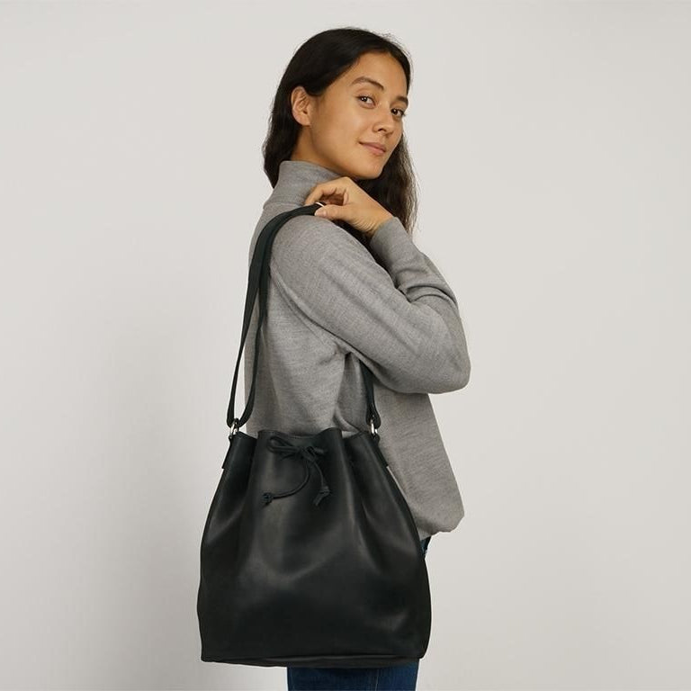 Eine Frau in einem grauen Pullover und blauen Jeans posiert seitlich mit einer schwarzen Lederumhängetasche und blickt vor einem schlichten weißen Hintergrund in die Kamera.