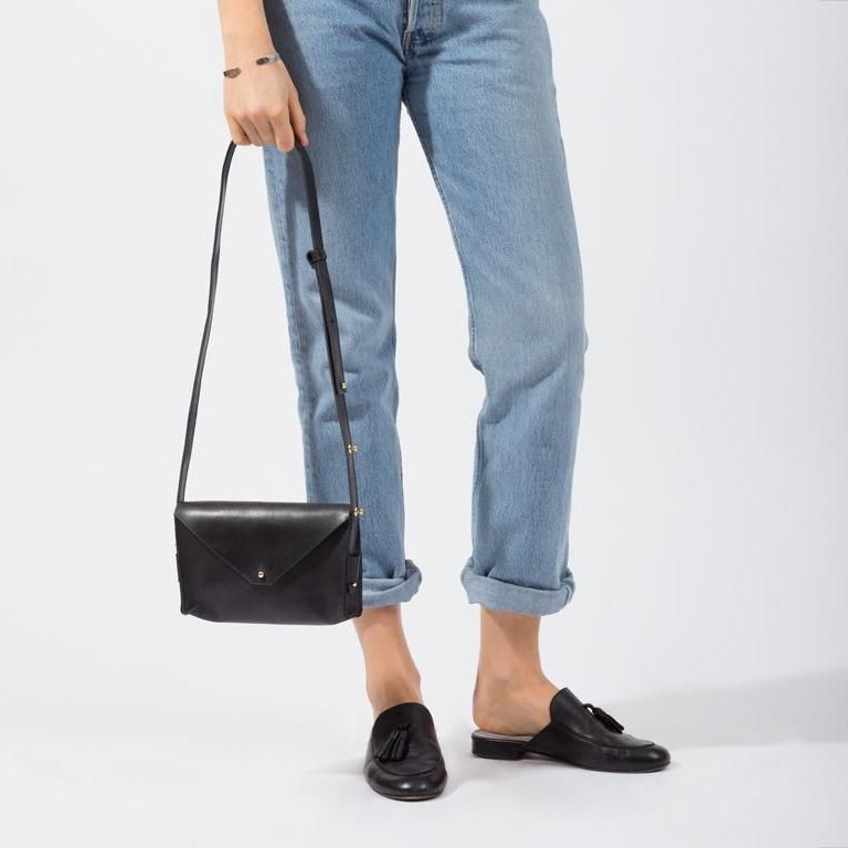 Eine Person in hellblauen, hochgekrempelten Jeans und schwarzen Halbschuhen hält eine schwarze Lederumhängetasche mit Überschlag und goldenem Schnappverschluss vor einem schlichten weißen Hintergrund.