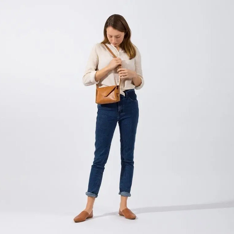 Eine Frau steht vor einem schlichten weißen Hintergrund und schaut in eine kleine braune Umhängetasche. Sie trägt eine helle Bluse, blaue, hochgekrempelte Jeans und braune, flache Schuhe.