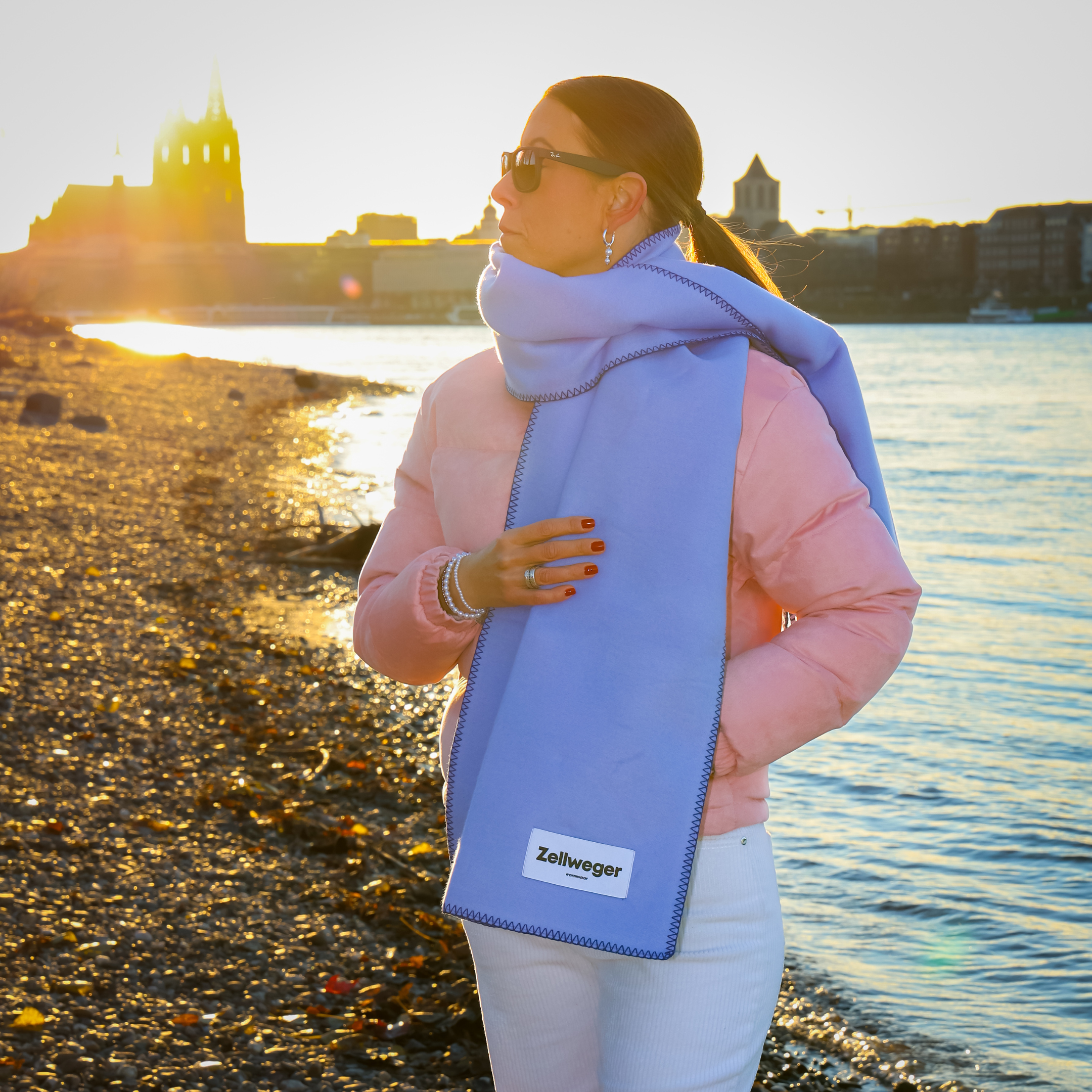 Frau in rosa Jacke mit blauem Schal steht am Flussufer bei Sonnenuntergang, trägt Sonnenbrille.