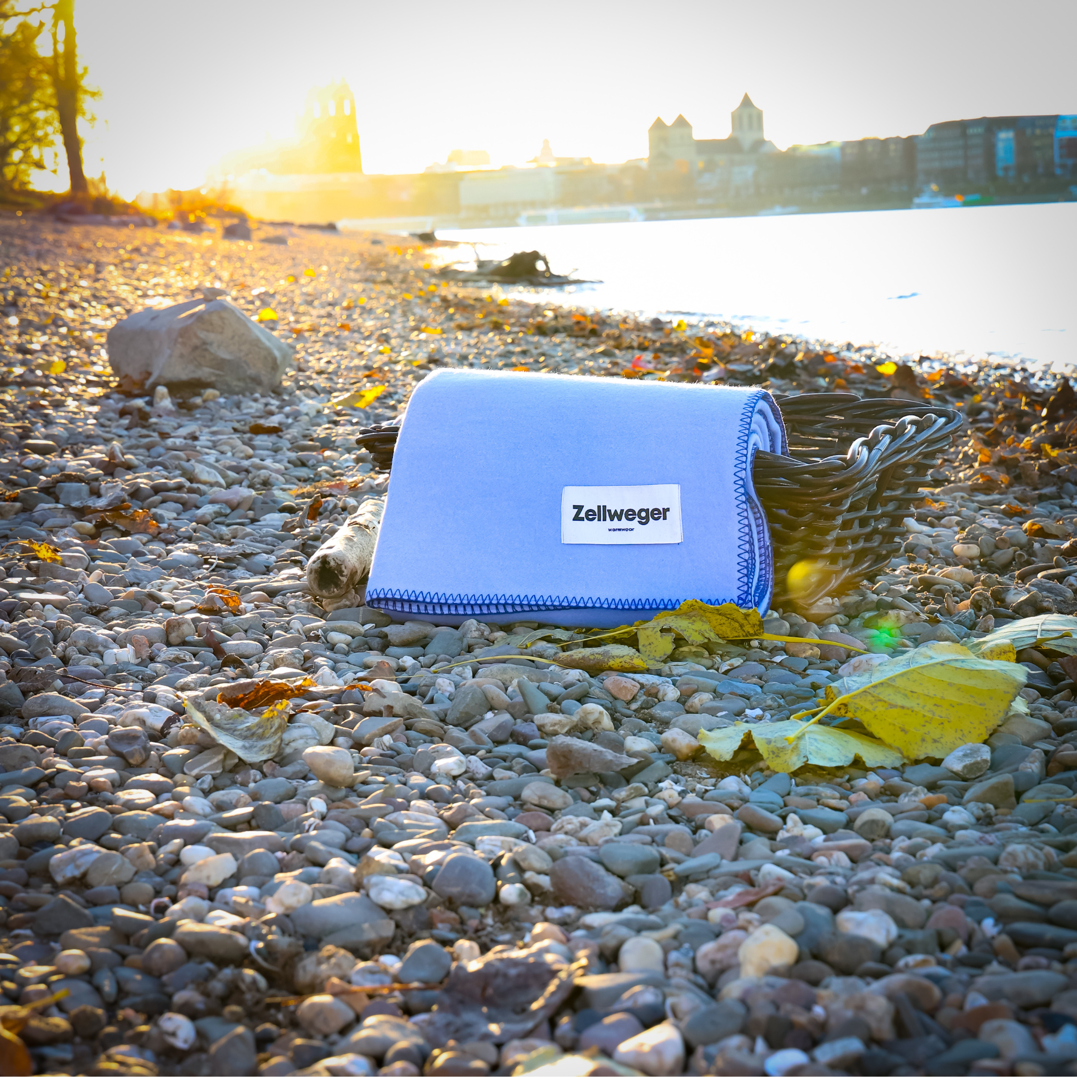Zusammengefaltete Decke mit "Zellweger"-Label liegt am kiesigen Flussufer bei Sonnenuntergang, Herbstblätter drumherum.