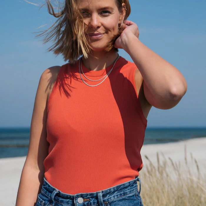 Eine Frau mit kurzen, hellbraunen Haaren, die ein orangefarbenes ärmelloses Oberteil und blaue Jeans trägt, steht an einem Strand, lächelt leicht und berührt mit einer Hand ihr Haar. Im Hintergrund sind das Meer und der Himmel zu sehen.
