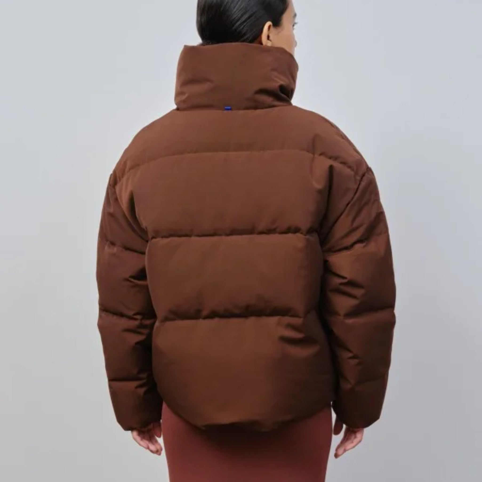 Person trägt eine braune, gepolsterte Jacke mit hohem Kragen, von hinten gesehen, vor neutralem Hintergrund.