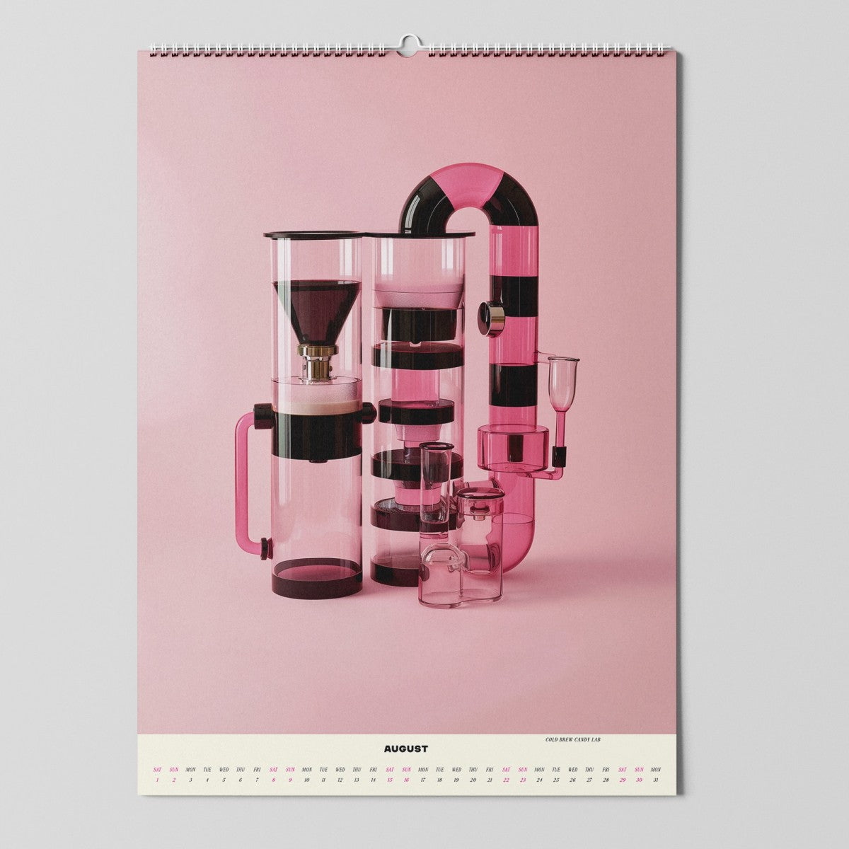 Ein Wandkalender, der bis August geöffnet ist, zeigt ein Foto von verschiedenen geometrischen, modernen Küchenbehältern und -geräten in klaren, schwarzen und rosafarbenen Farben, die vor einem rosa Hintergrund angeordnet sind.