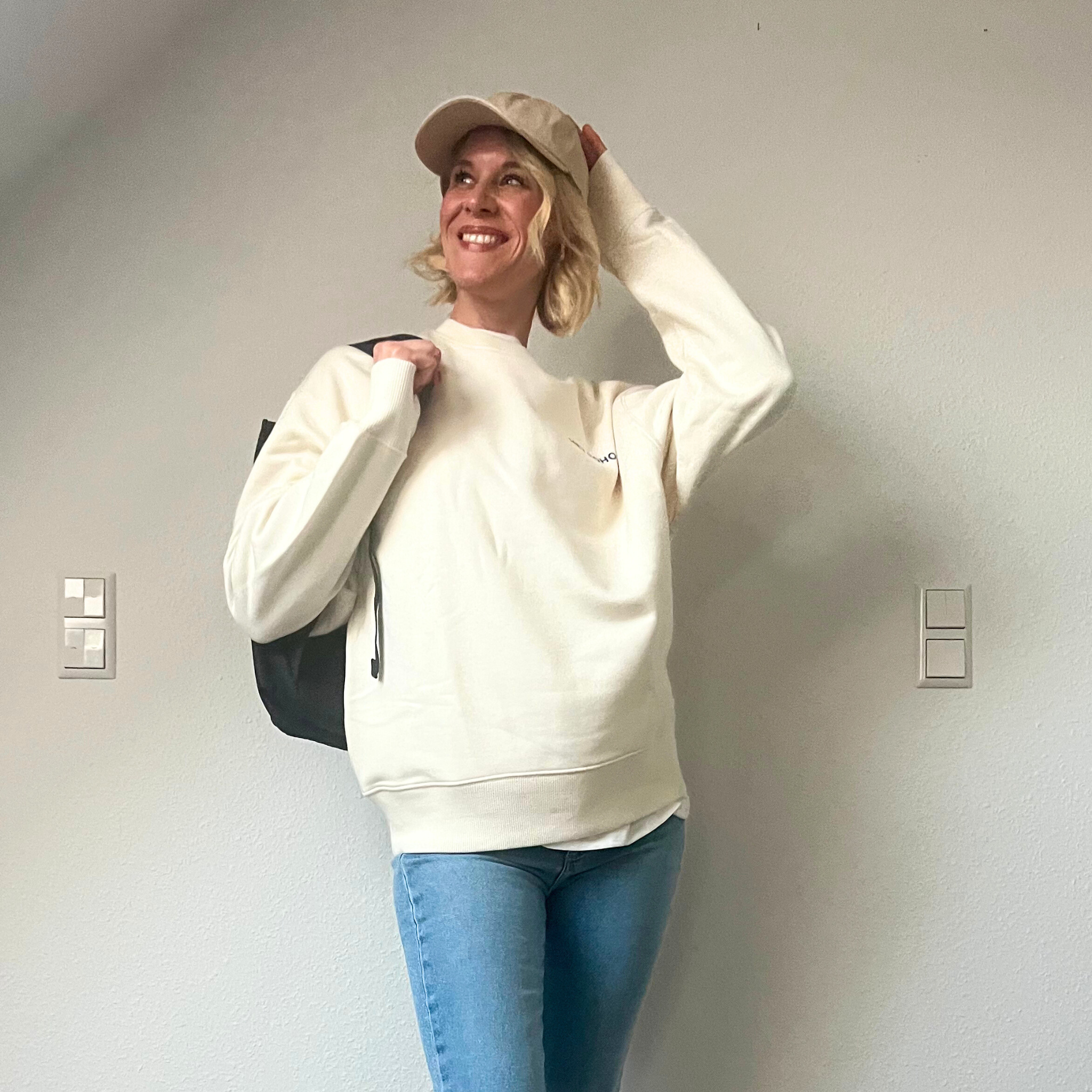 Frau in beigem Pullover und Jeans steht lächelnd vor einer Wand, mit einer Mütze und einem schwarzen Rucksack.