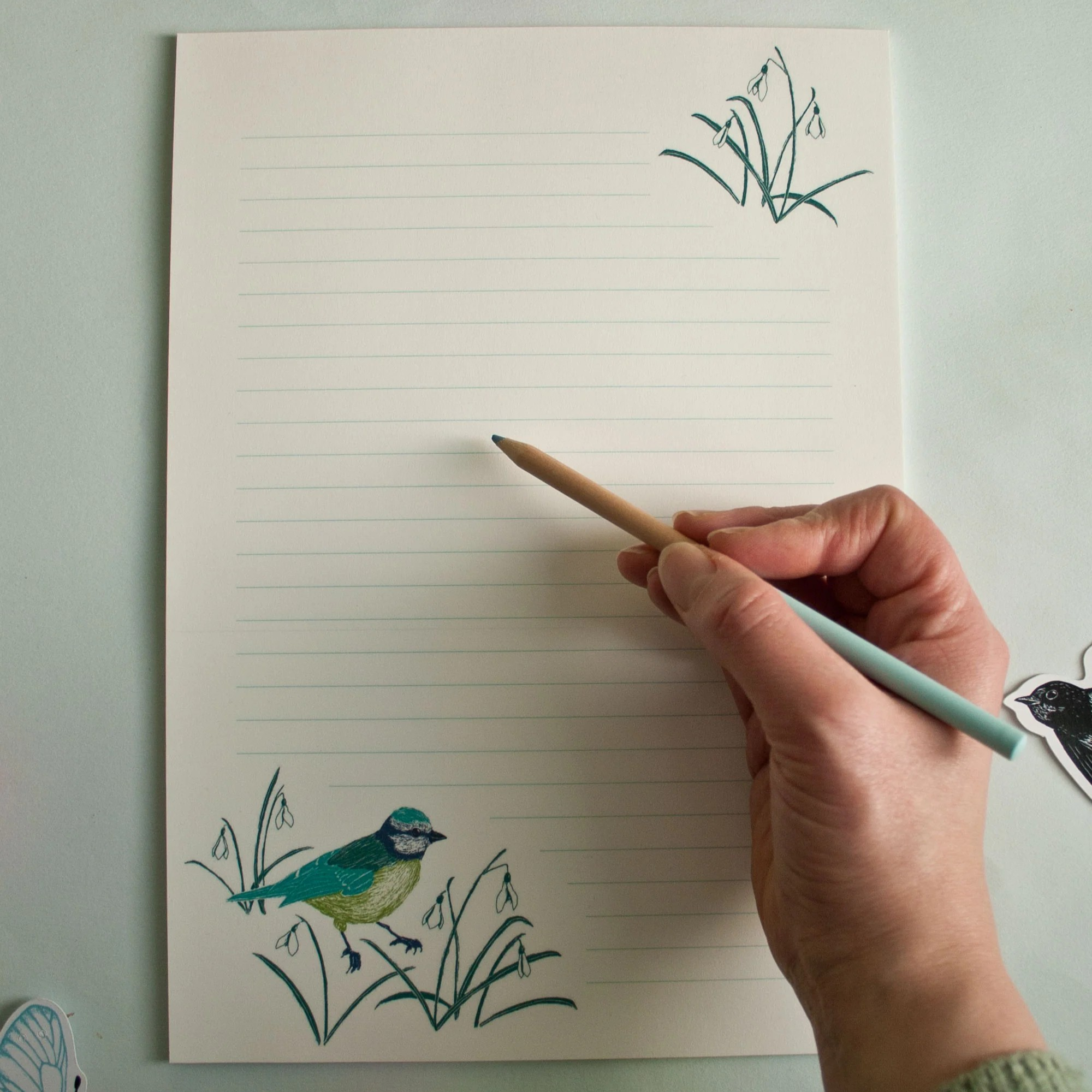 Hand schreibt mit einem Bleistift auf liniertes Papier mit gezeichnetem Vogel und Pflanzenmotiv in den Ecken.