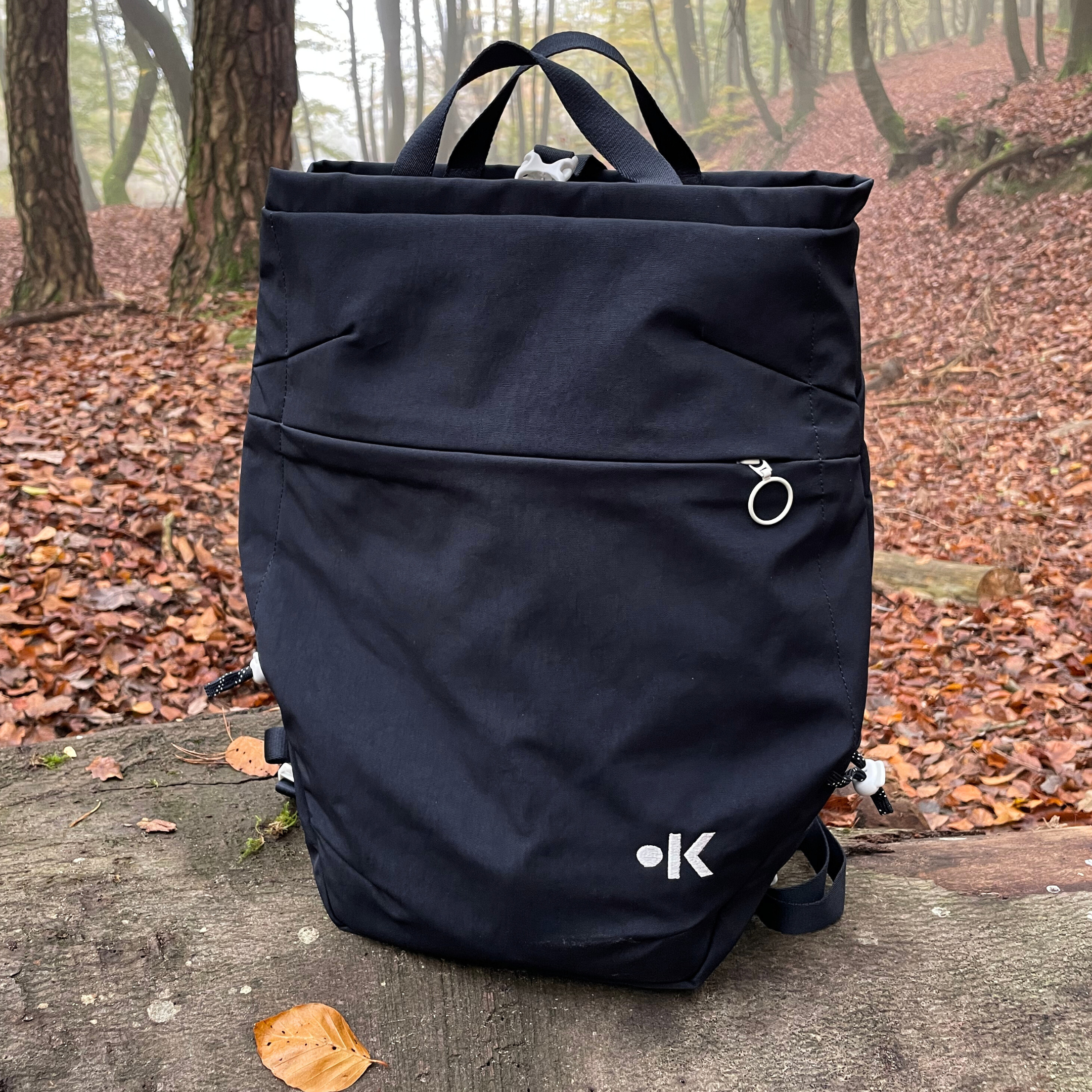 Schwarzer Rucksack auf Baumstumpf in herbstlichem Wald mit braunen Blättern am Boden.