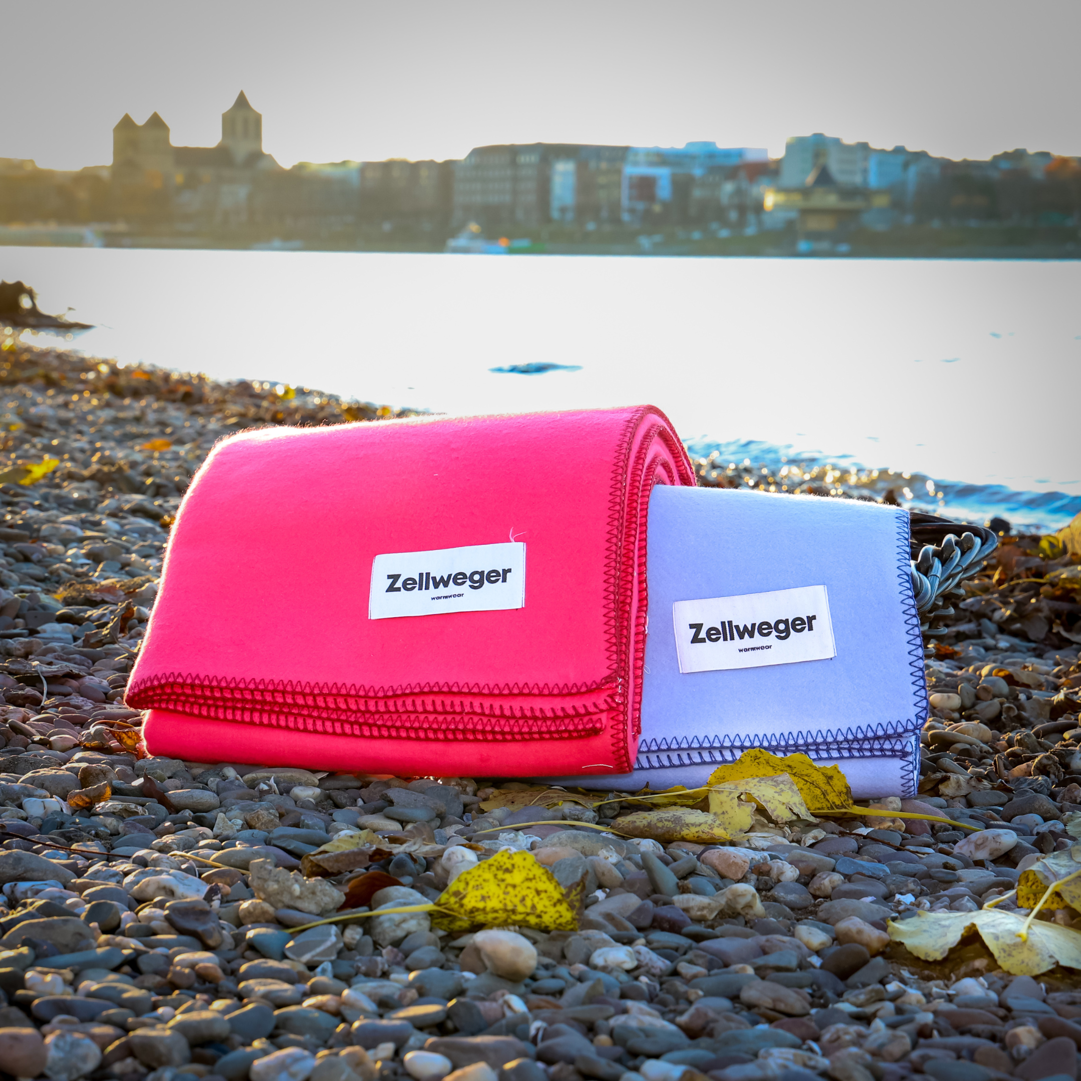 Zwei Zellweger-Decken, eine rosa und eine graue, liegen auf einem Kiesstrand mit Stadtansicht im Hintergrund.