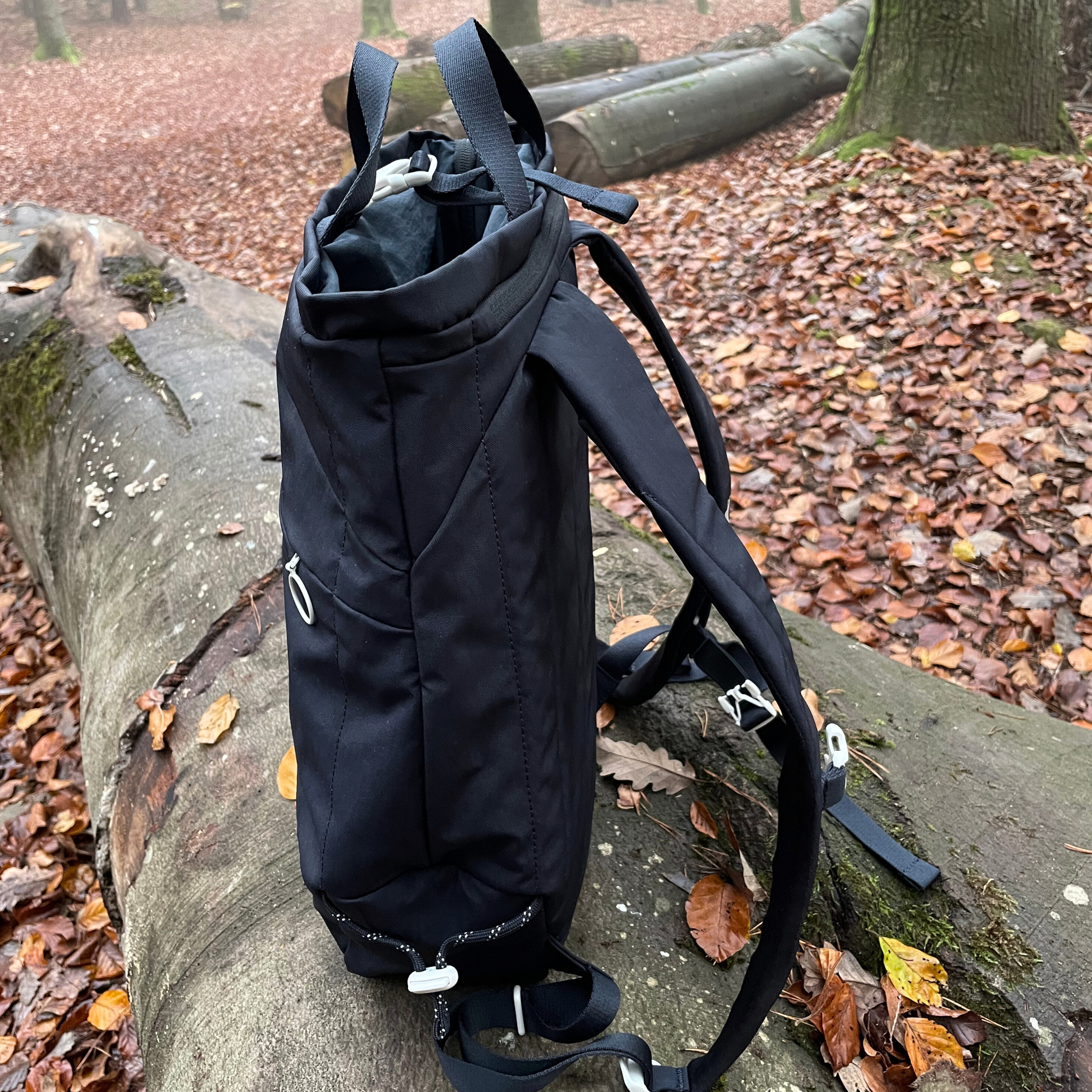 Schwarzer Rucksack auf einem gefallenen Baumstamm im herbstlichen Wald voller Laub.